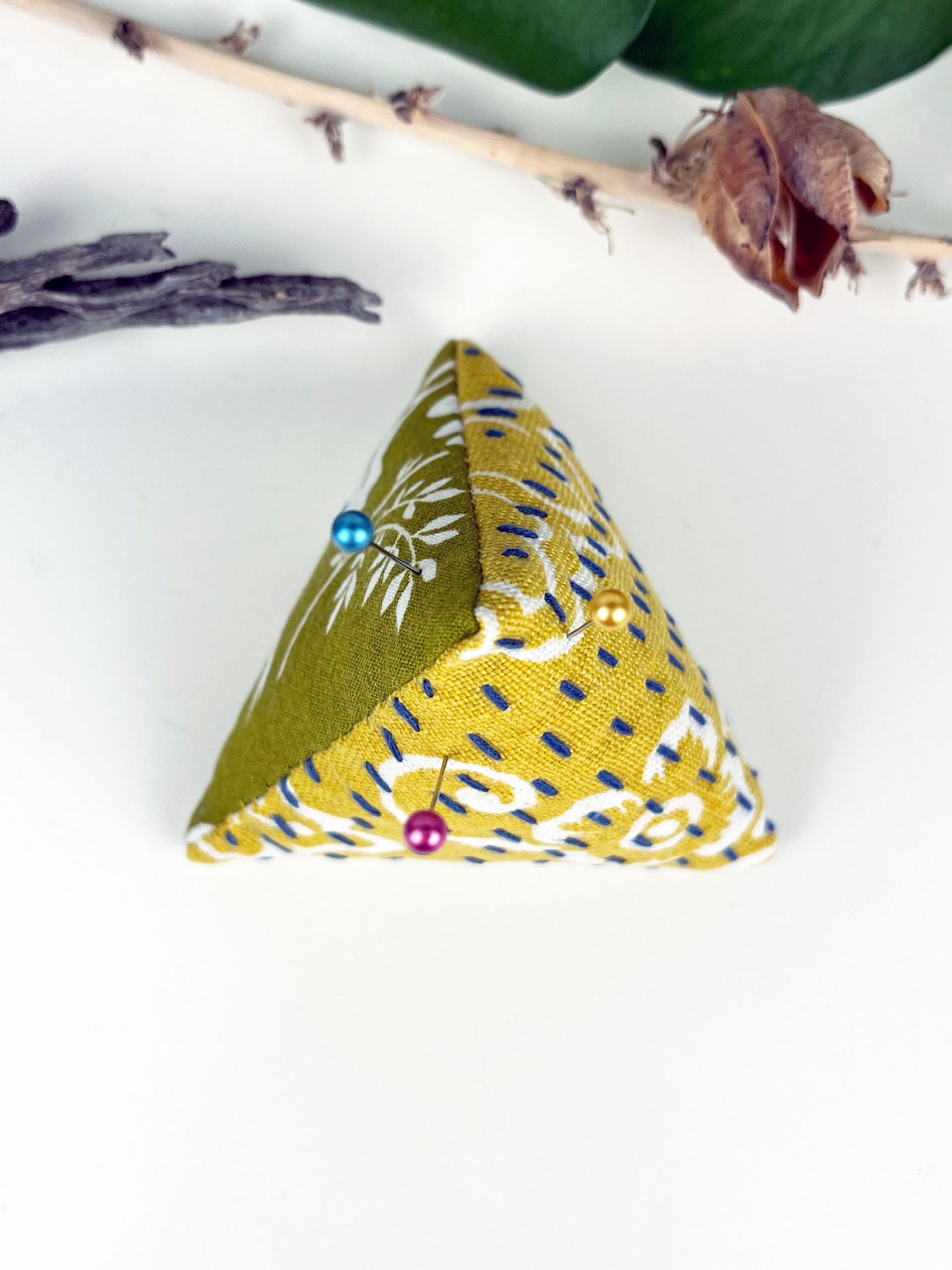 birds eye view of a pyramid pincushion made from floral olive and mustard fabric with contrasting sashiko stitches on a white counter with dried twigs seedpods and a pothos plant in the background