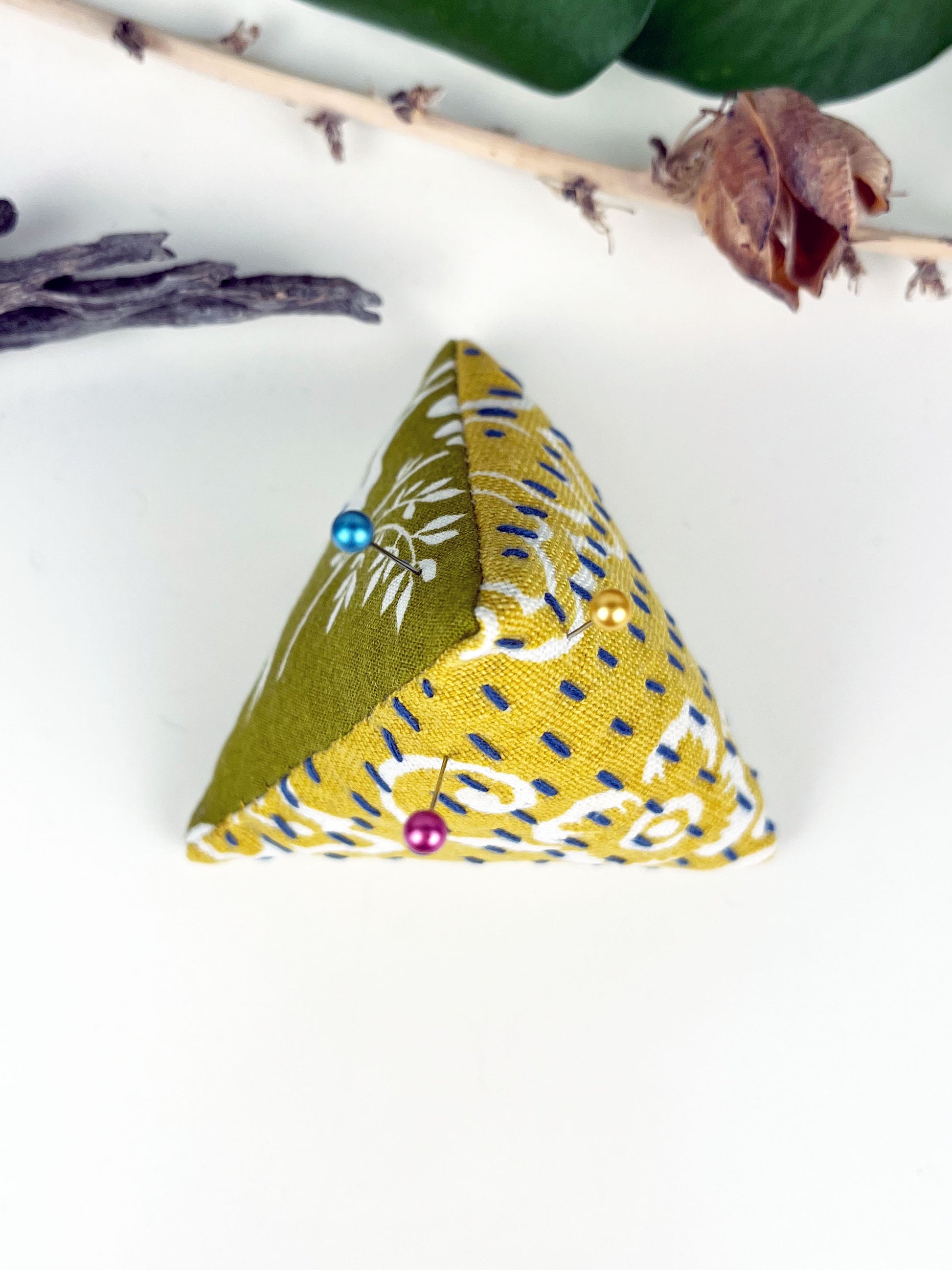 birds eye view of a pyramid pincushion made from floral olive and mustard fabric with contrasting sashiko stitches on a white counter with dried twigs seedpods and a pothos plant in the background