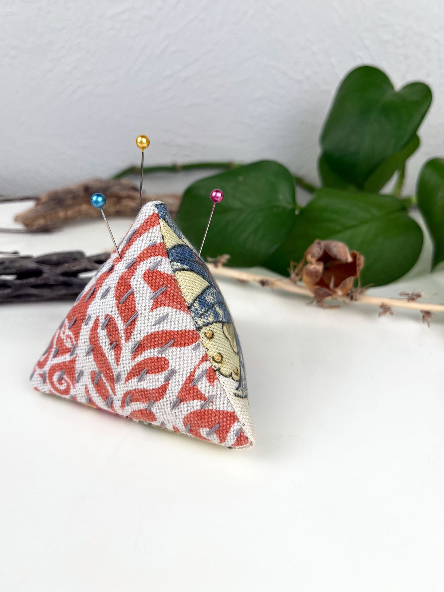 a pyramid pincushion made from floral and orange upholstery fabric with contrasting sashiko stitches on a white counter with dried twigs seedpods and a pothos plant in the background