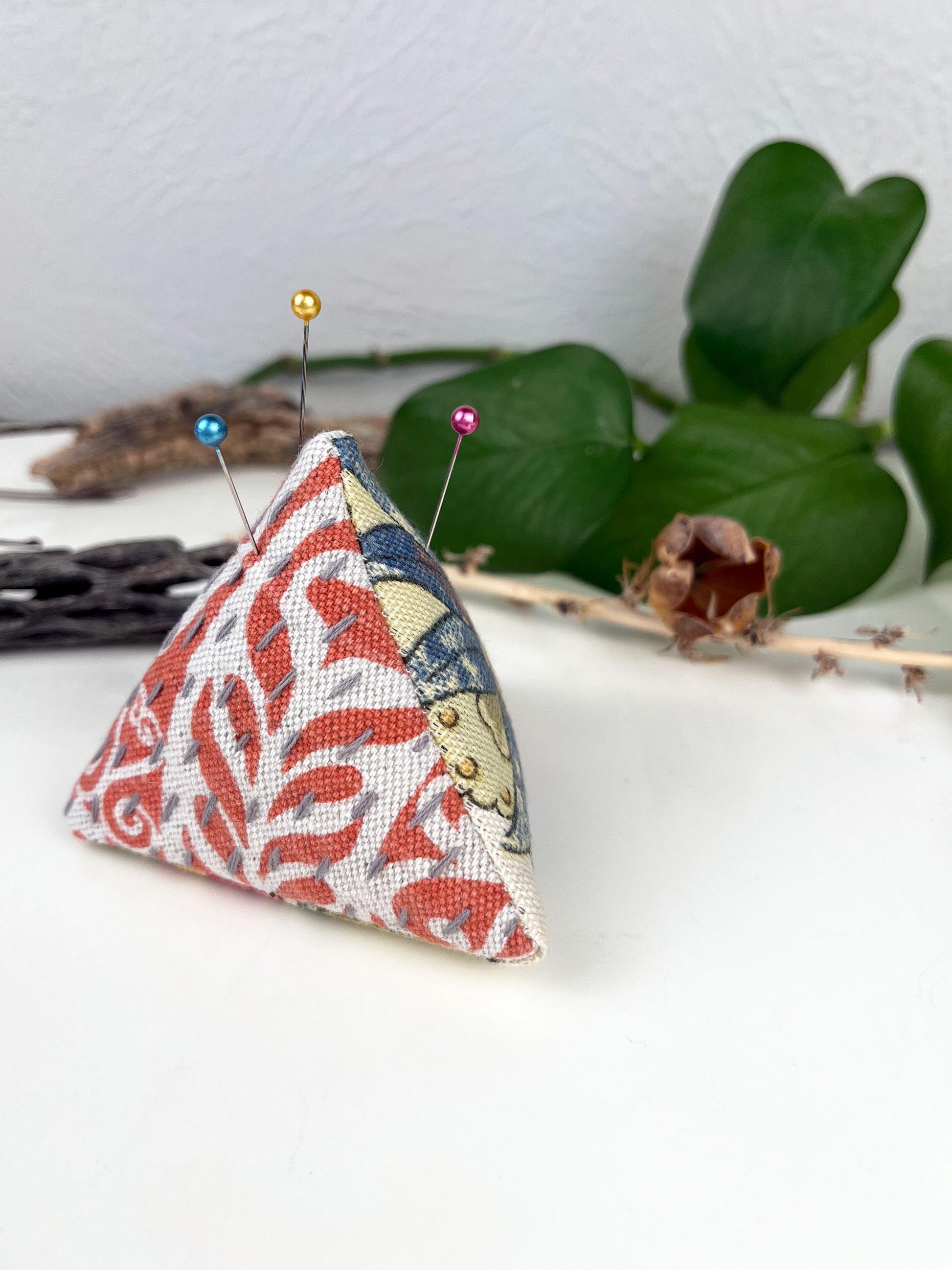 a pyramid pincushion made from floral and orange upholstery fabric with contrasting sashiko stitches on a white counter with dried twigs seedpods and a pothos plant in the background