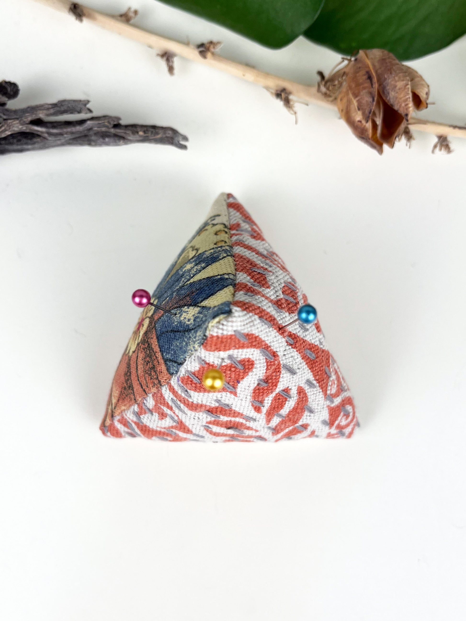birds eye view of a pyramid pincushion made from floral and orange upholstery fabric with contrasting sashiko stitches on a white counter with dried twigs seedpods and a pothos plant in the background