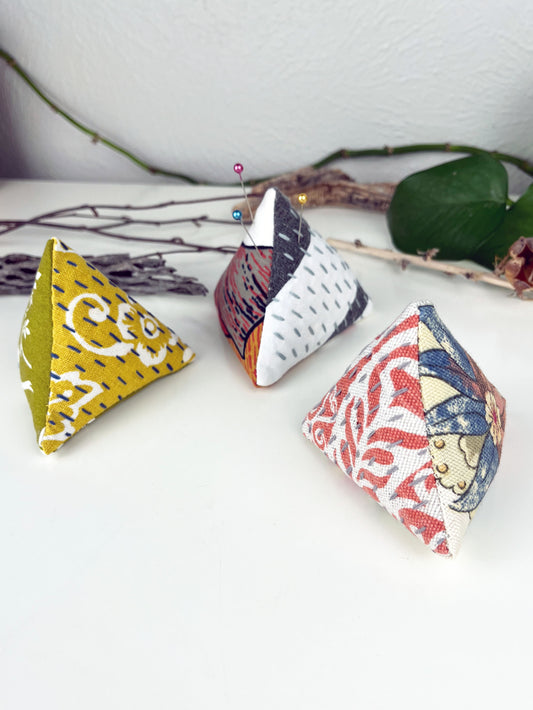 a group of three pyramid shaped pincushion made from a variety of colorful  fabrics with contrasting sashiko stitches on a white counter with dried twigs cactus seedpods and a pothos plant in the background
