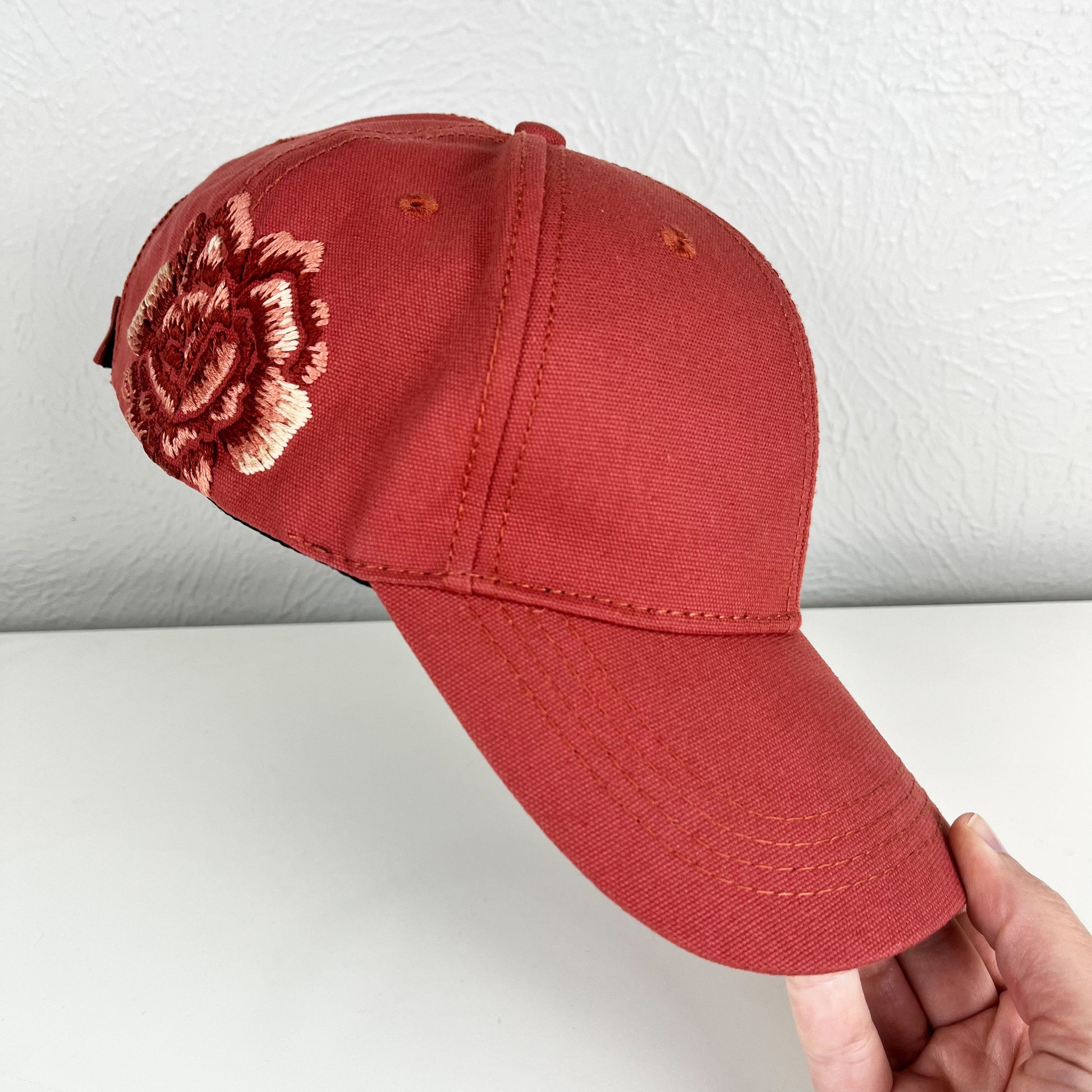 terra cotta colored baseball hat with a large hand embroidered peony on the side, also in shades of terra cotta, held up in the air at an angle