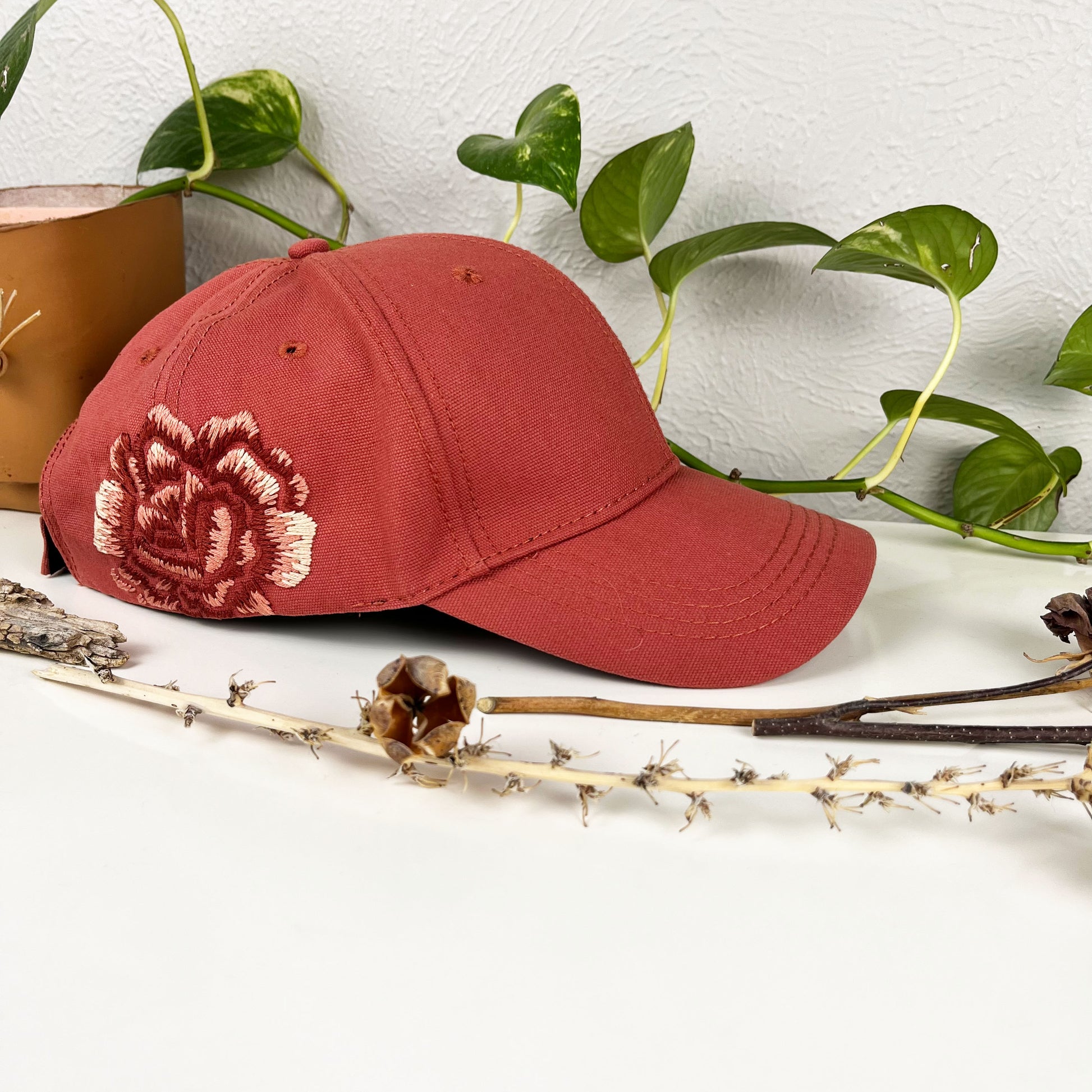 terra cotta colored baseball hat with a large hand embroidered peony on the side, also in shades of terra cotta, sitting on a dresser with a plant vine and dried flowers around it