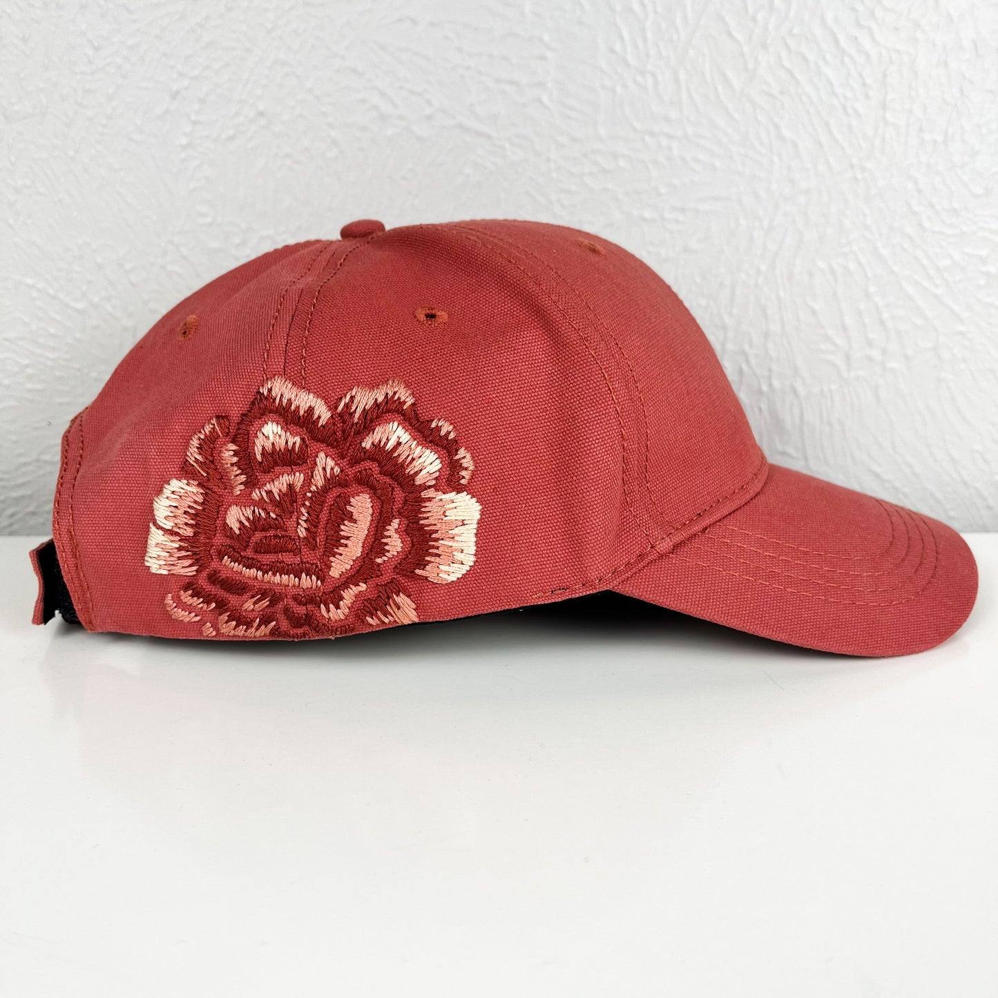 side view of a terra cotta colored baseball hat with a large hand embroidered peony on the side, also in shades of terra cotta, sitting on a white dresser