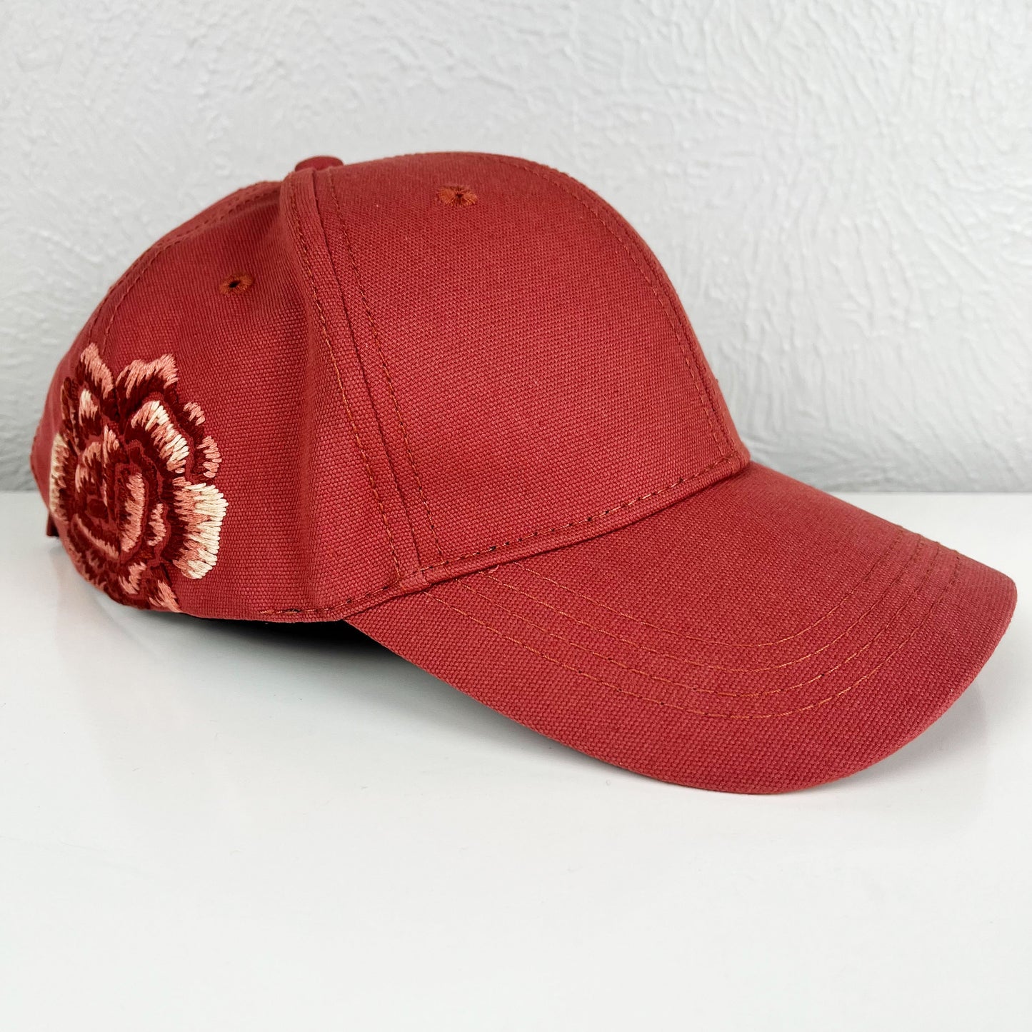 terra cotta colored baseball hat with a large hand embroidered peony on the side, also in shades of terra cotta, sitting at an angle on a white dresser