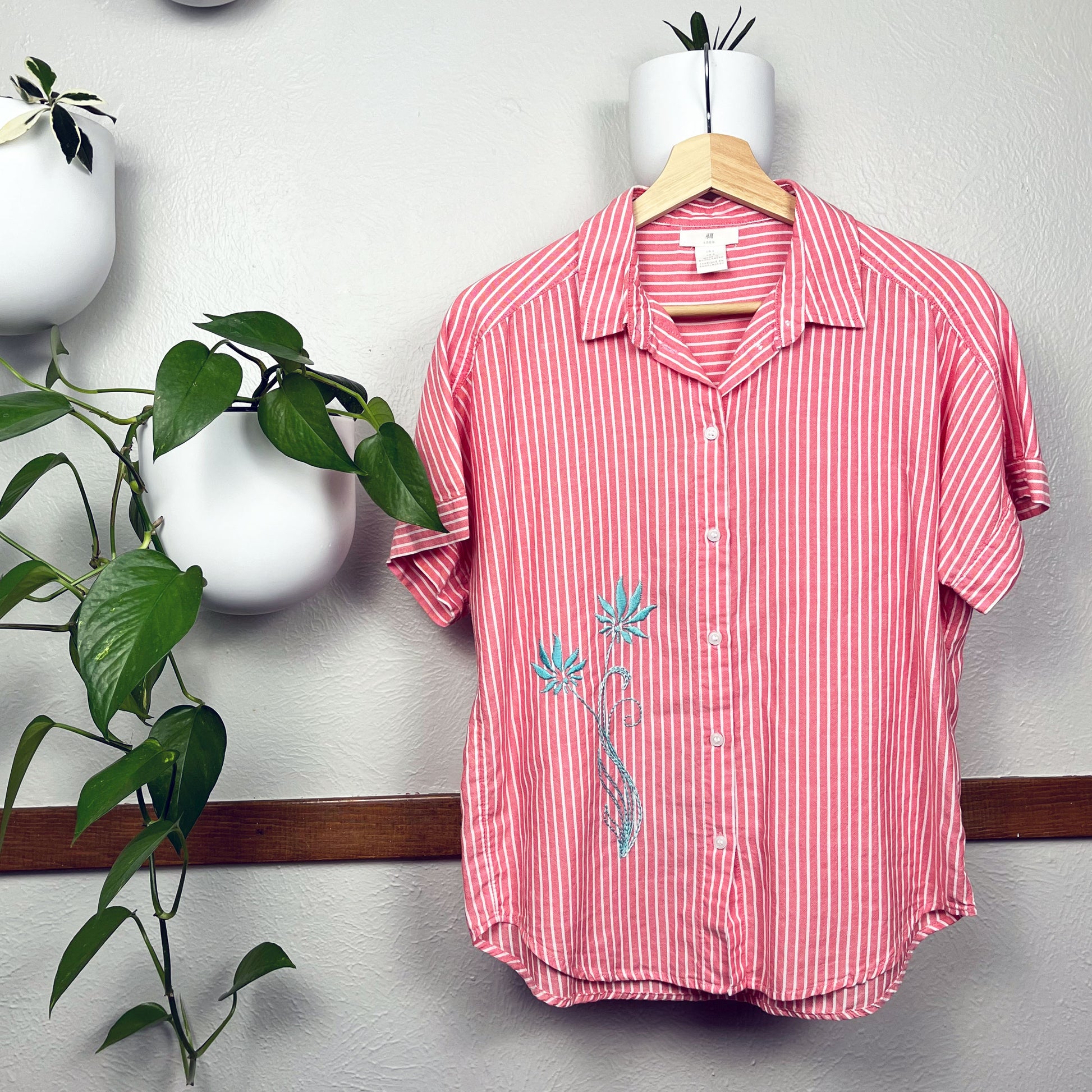 a red and white striped button-up short sleeve shirt, with two hand embroidered long stemmed flowers in aqua on the lower front right side of the shirt, on a wooden hanger with plants in round white pots hanging on the wall around it