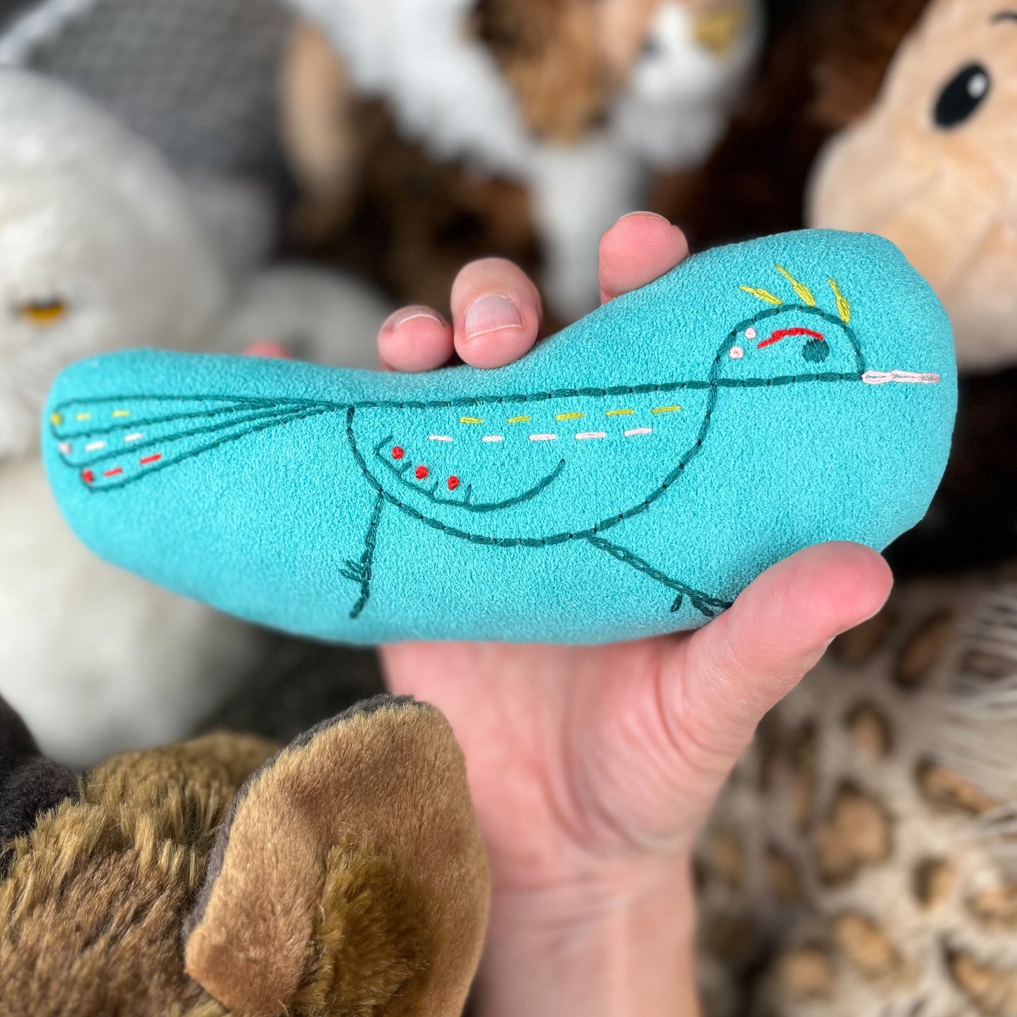a colorfully hand embroidered stuffed pillow animal of a roadrunner in seafoam green fabric, held in a hand with out-of-focus stuffed animals in the background