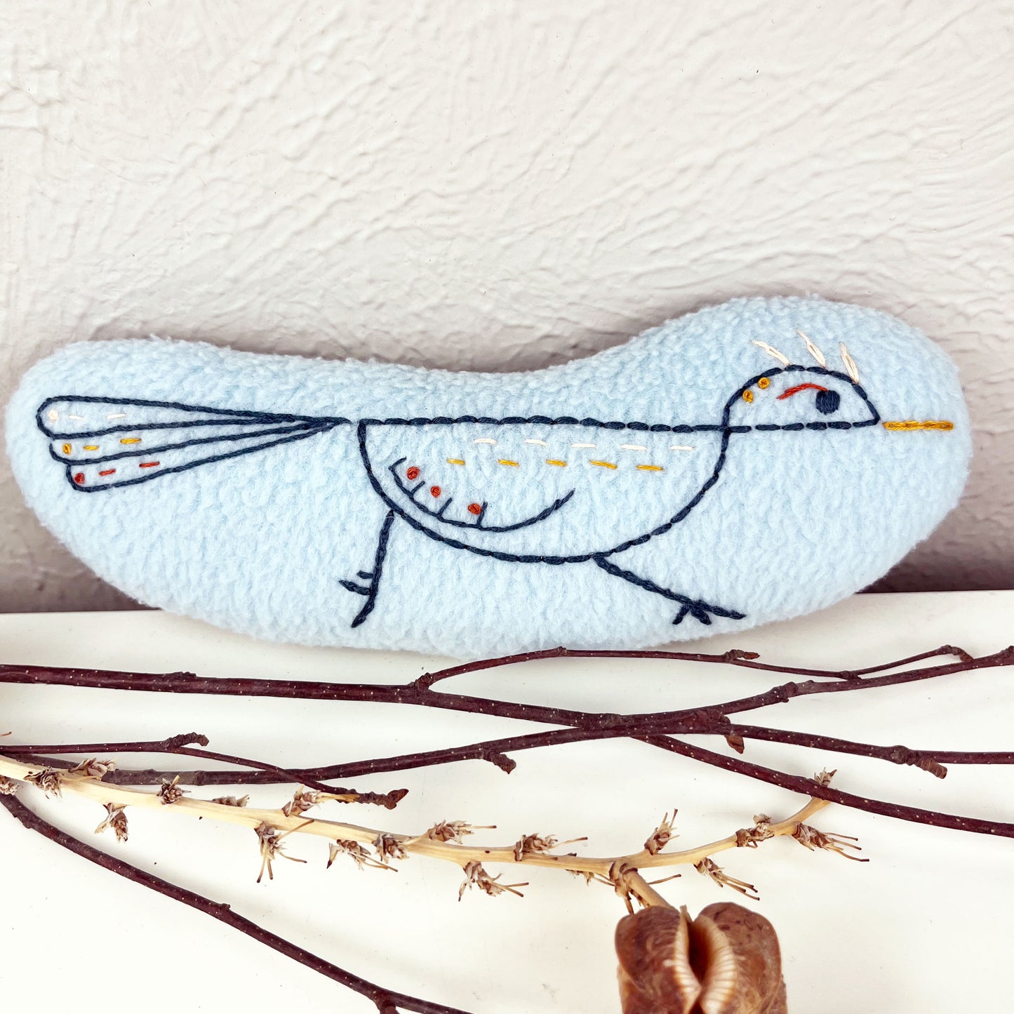 a colorfully hand embroidered stuffed pillow roadrunner on light blue fleece, sitting on a white counter with twigs and dried seed pods