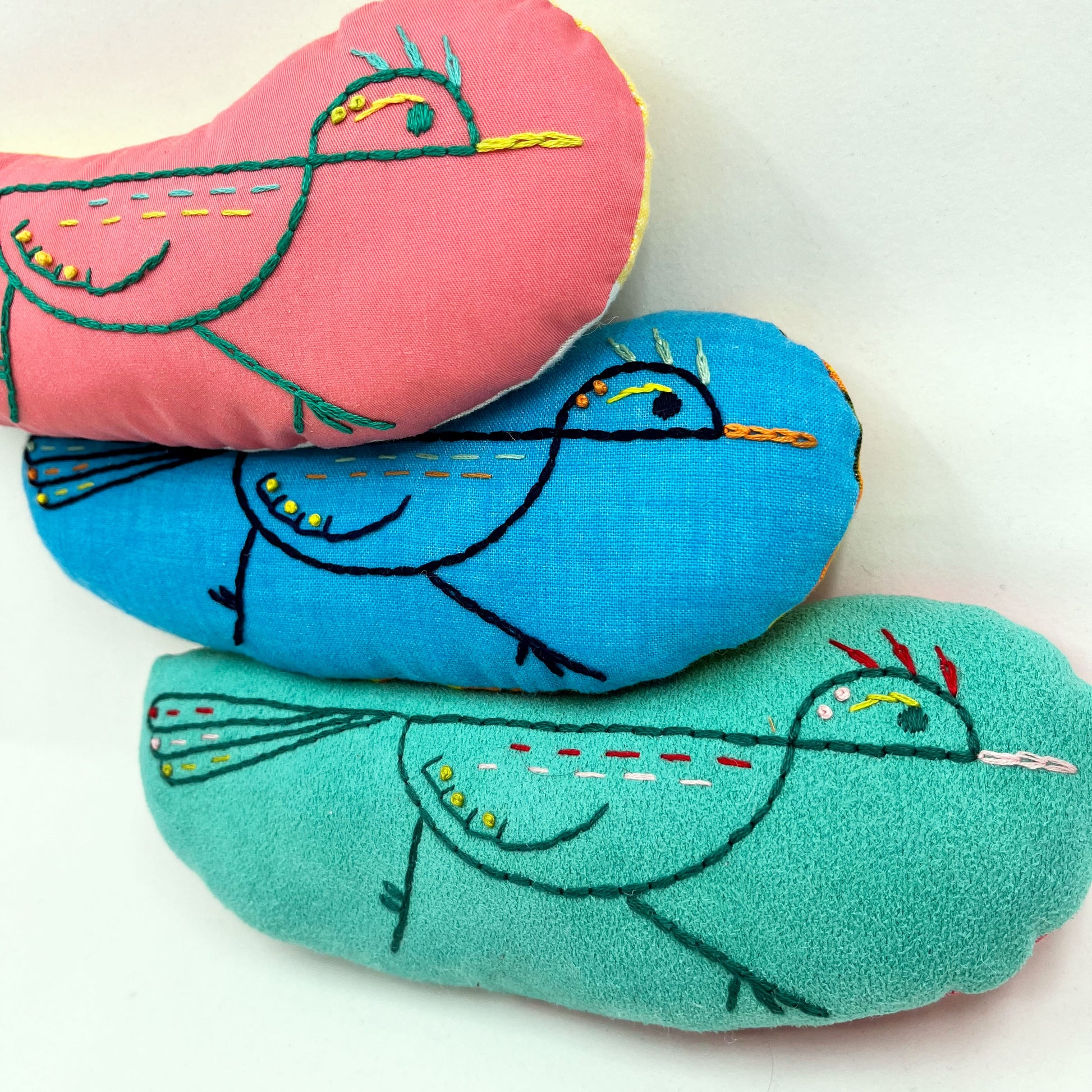 a group of colorfully hand embroidered roadrunner stuffed animal pillows in blue, seafoam green and coral fabric, in a pile on a white background