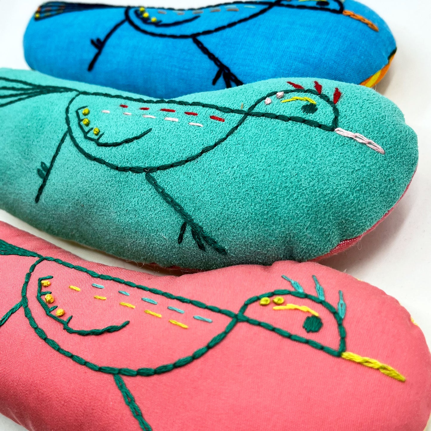 close up angled view of a group of colorfully hand embroidered roadrunner stuffed animal pillows in blue, seafoam green and coral fabric, in a pile on a white background