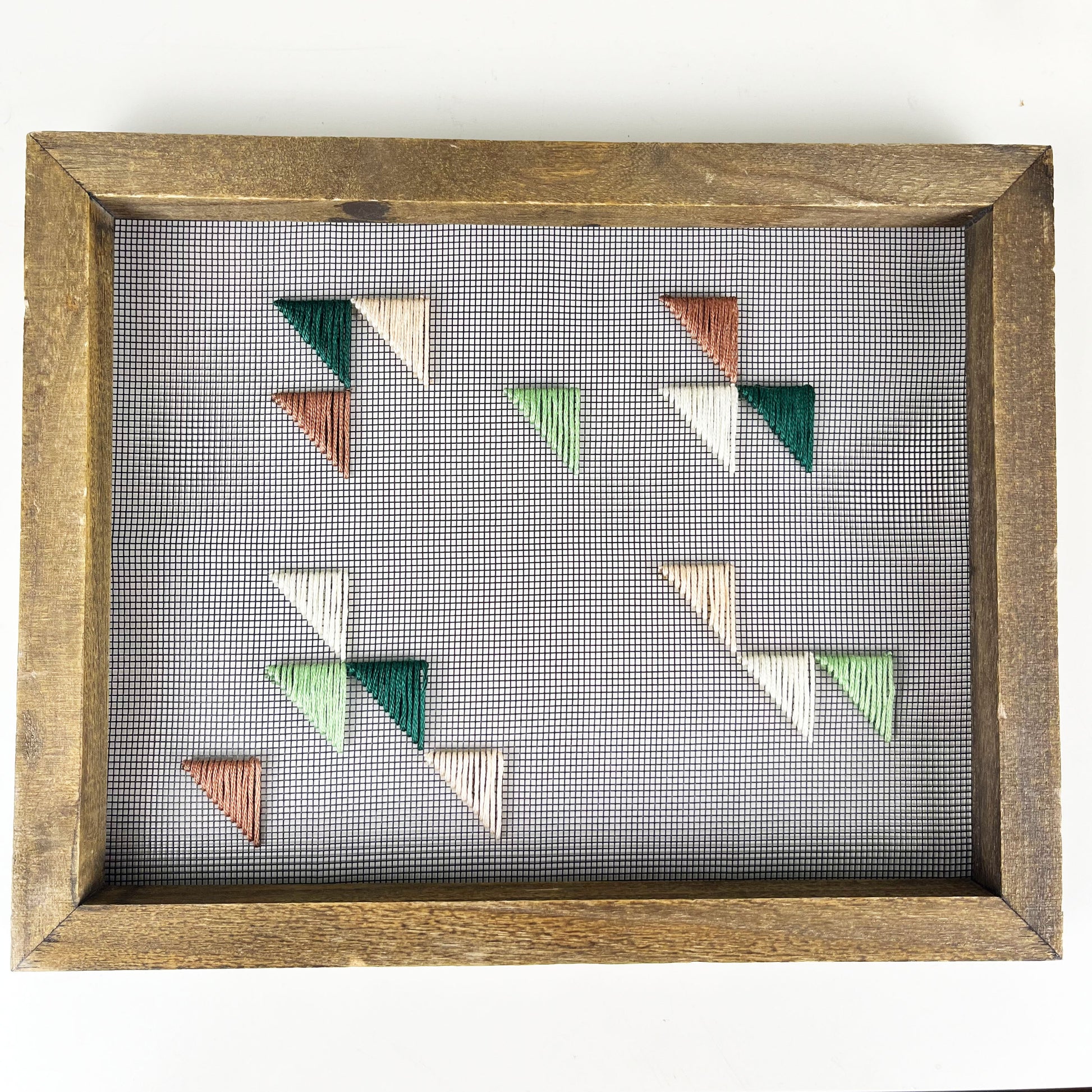 a piece of window screen hand stitched with green mauve peach and ivory triangles in an abstract design, in a wood frame