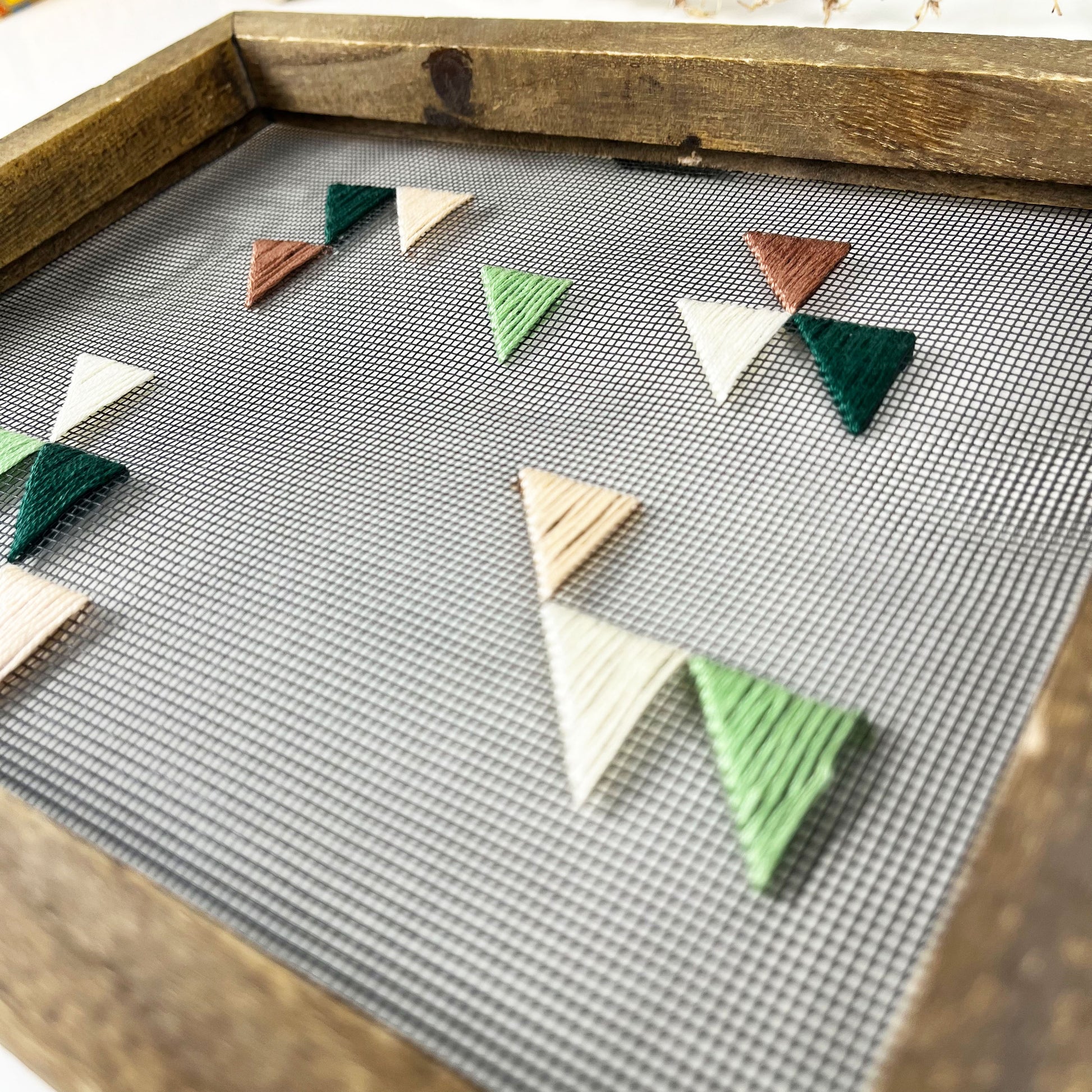 close up angled view of a piece of window screen hand stitched with green mauve peach and ivory triangles in an abstract design, in a wood frame