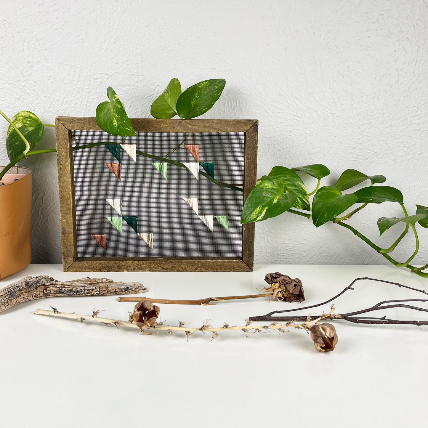 a piece of window screen hand stitched with green mauve peach and ivory triangles in an abstract design, in a wood frame, on a white counter, with a pothos behind it and sticks/dried flowers in front of it