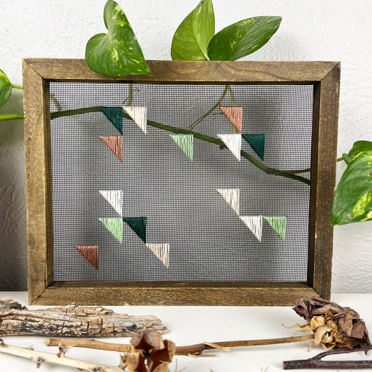 a piece of window screen hand stitched with green mauve peach and ivory triangles in an abstract design, in a wood frame, on a white counter, with a pothos behind it and sticks/dried flowers in front of it