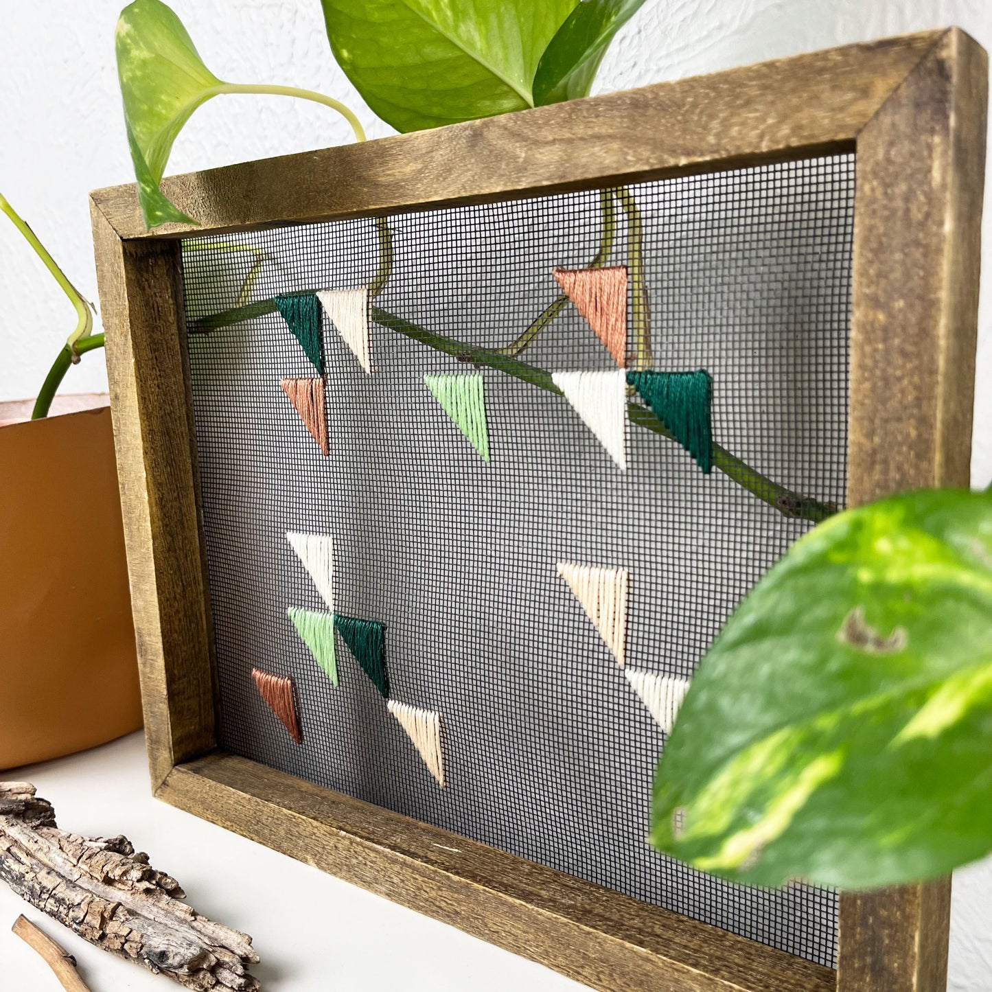close up angled view of a piece of window screen hand stitched with green mauve peach and ivory triangles in an abstract design, in a wood frame, on a white counter, with a pothos behind it and sticks/dried flowers in front of it
