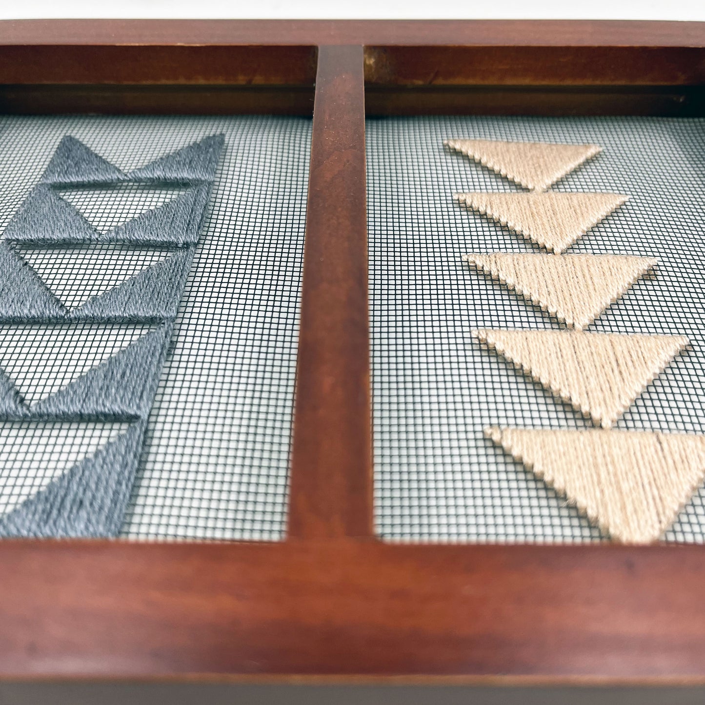 Small double panel wood frame holding window screen hand stitched with peach and grey triangles that form shapes of flying geese quilt pattern, on the left side a column of the border triangles in grey, on the right side the center triangles in peach