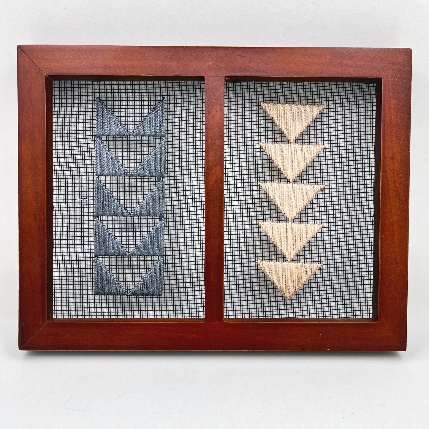 Small double panel wood frame holding window screen hand stitched with peach and grey triangles that form shapes of flying geese quilt pattern, on the left side a column of the border triangles in grey, on the right side the center triangles in peach