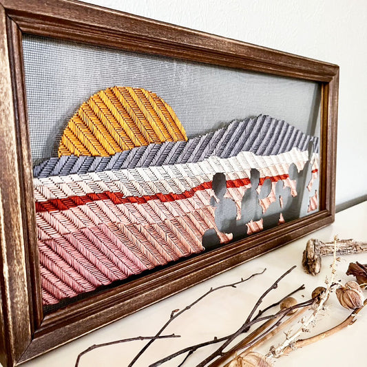 old window screen in a brown wood frame hand stitched with a landscape image of a mountain, small volcanoes and a setting sun, with prickly pears in negative space in the foreground