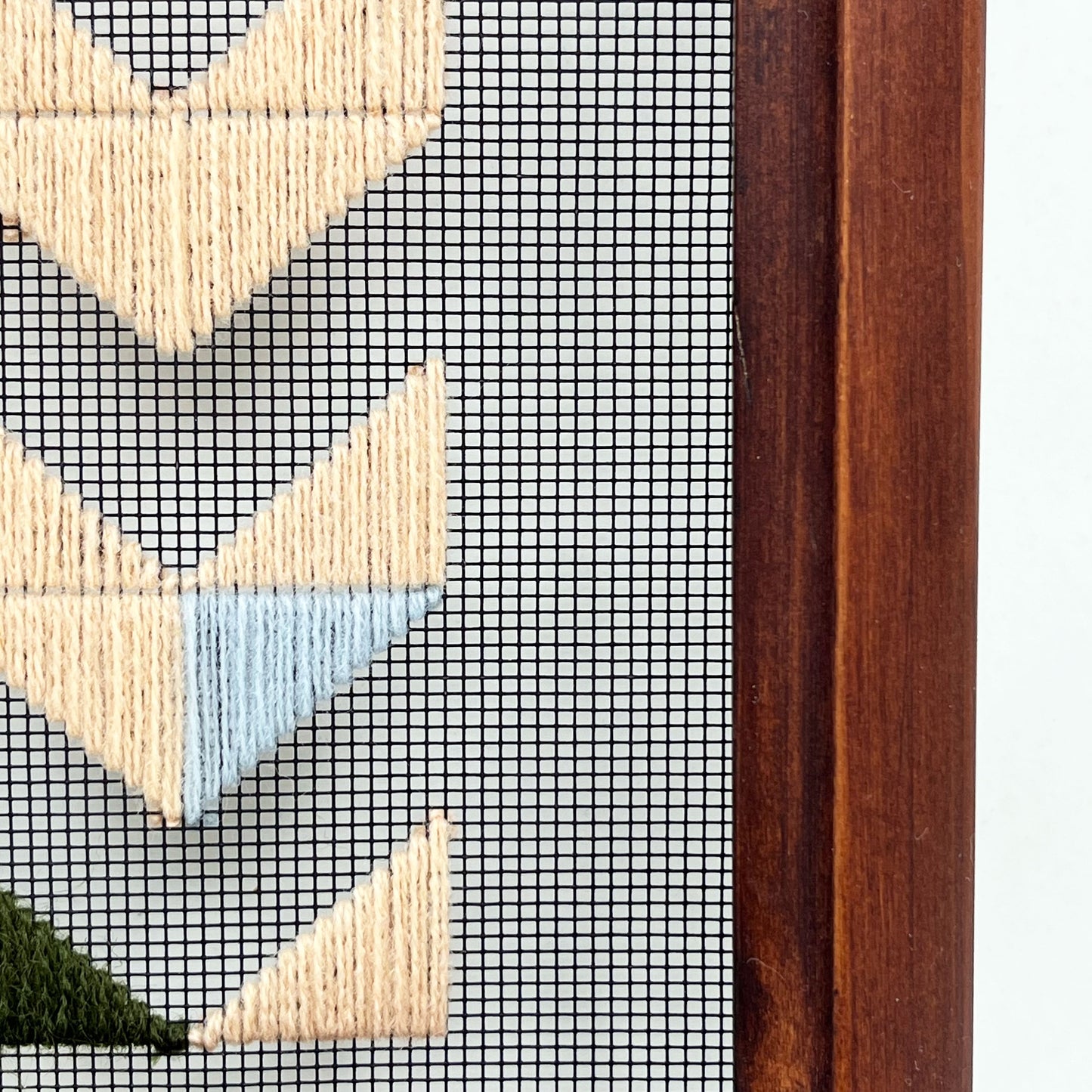 Close up detail of a small wood frame holding window screen hand stitched with triangles forming the shapes of chevrons disappearing off the bottom edge in mostly peach embroidery floss, with accents of light blue and olive green