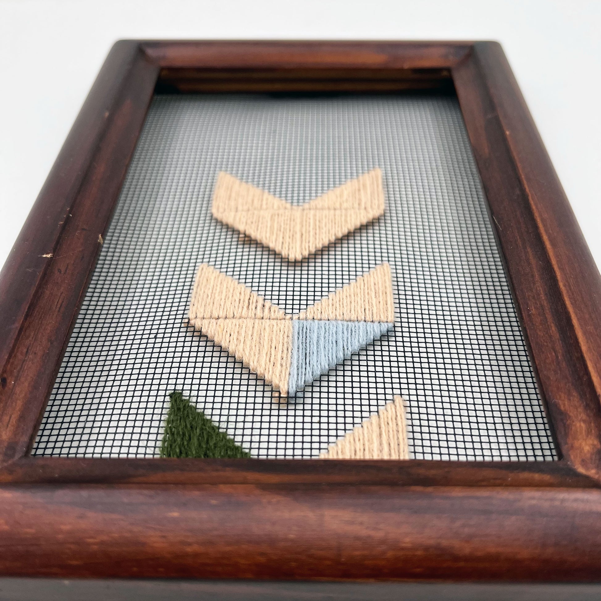 Close up detail angled perspective of a small wood frame holding window screen hand stitched with triangles forming the shapes of chevrons disappearing off the bottom edge in mostly peach embroidery floss, with accents of light blue and olive green