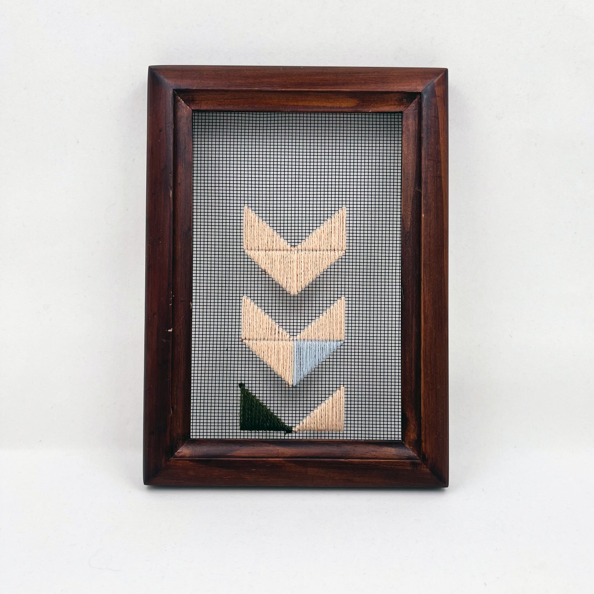 a small wood frame holding window screen hand stitched with triangles forming the shapes of chevrons disappearing off the bottom edge in mostly peach embroidery floss, with accents of light blue and olive green