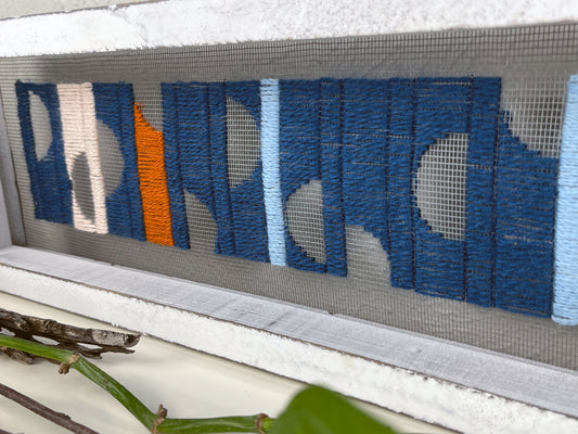 close up view of a window screen framed in a long thick white frame, the screen is hand stitched with columns of midnight blue light blue rust and peach with negative space semi circles, on a white counter with pothos vines and dried sticks around it