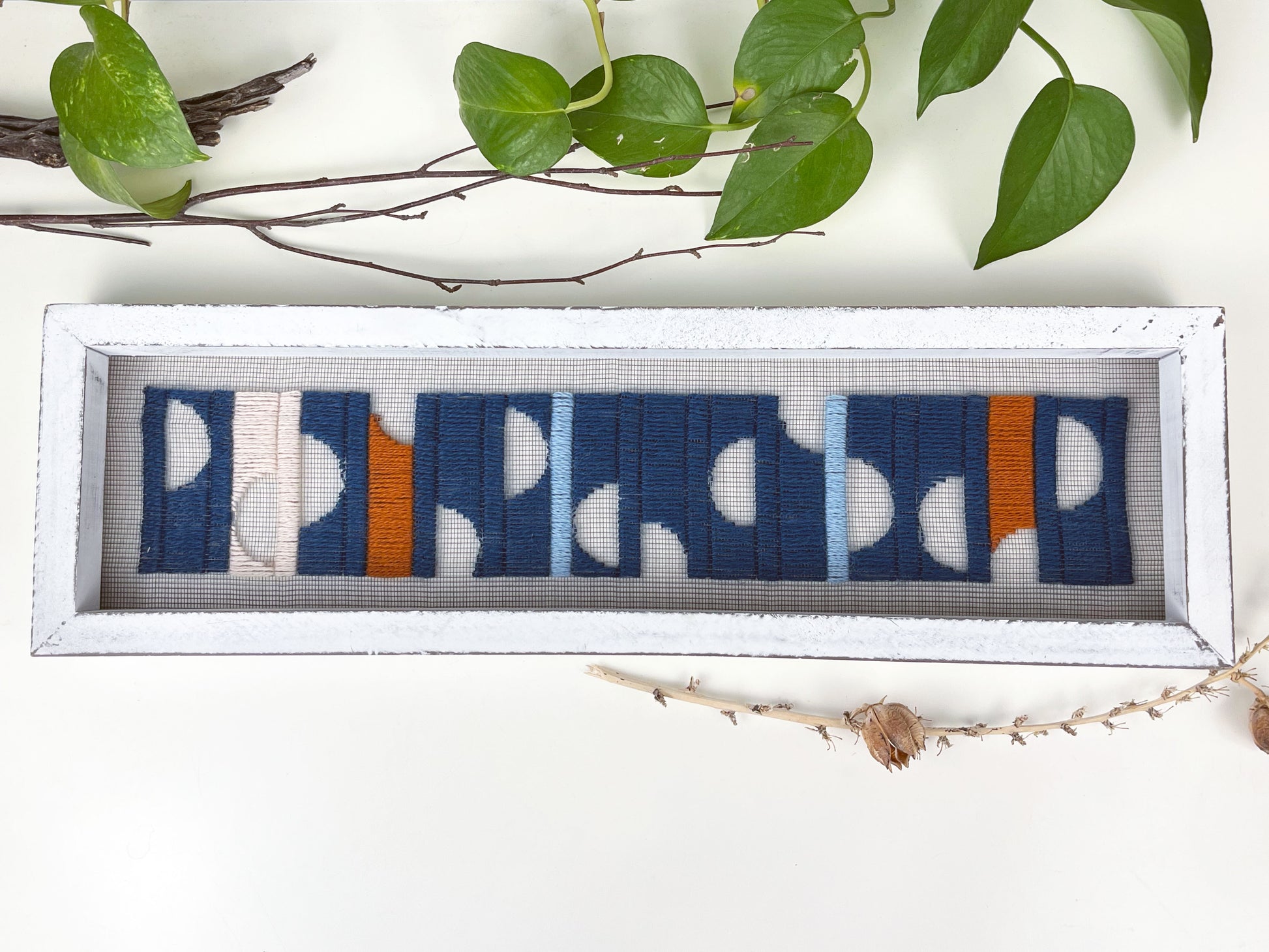 a window screen framed in a long thick white frame, the screen is hand stitched with columns of midnight blue light blue rust and peach with negative space semi circles, on a white counter with pothos vines and dried sticks around it