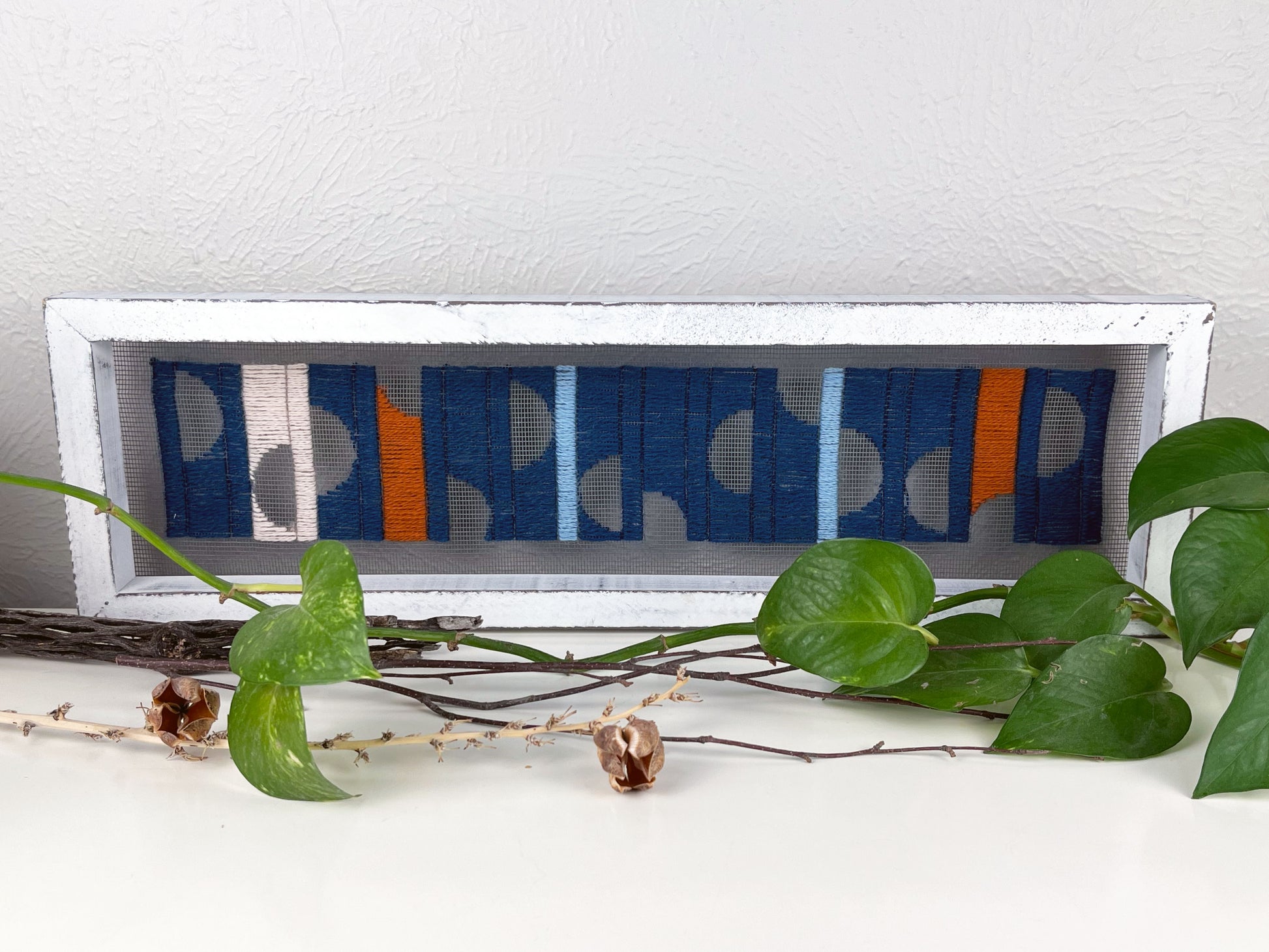 close up view of a window screen framed in a long thick white frame, the screen is hand stitched with columns of midnight blue light blue rust and peach with negative space semi circles, on a white counter with pothos vines and dried sticks around it