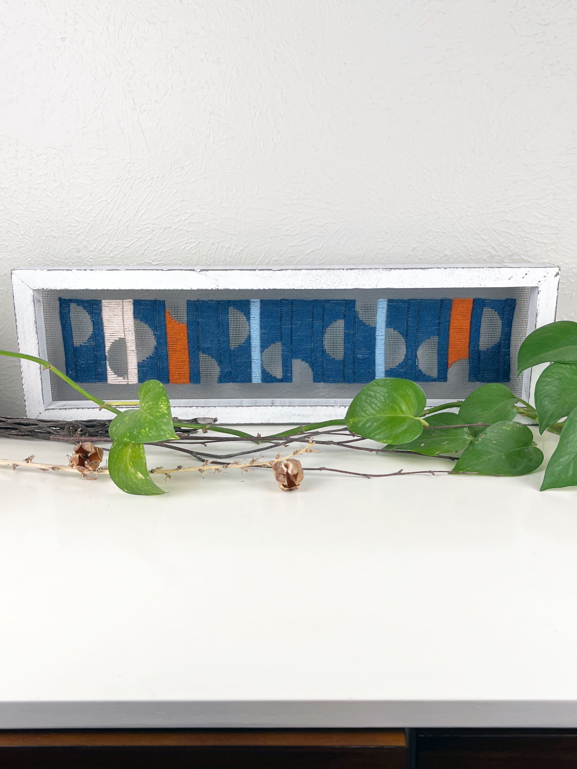 a window screen framed in a long thick white frame, the screen is hand stitched with columns of midnight blue light blue rust and peach with negative space semi circles, on a white counter with pothos vines and dried sticks around it