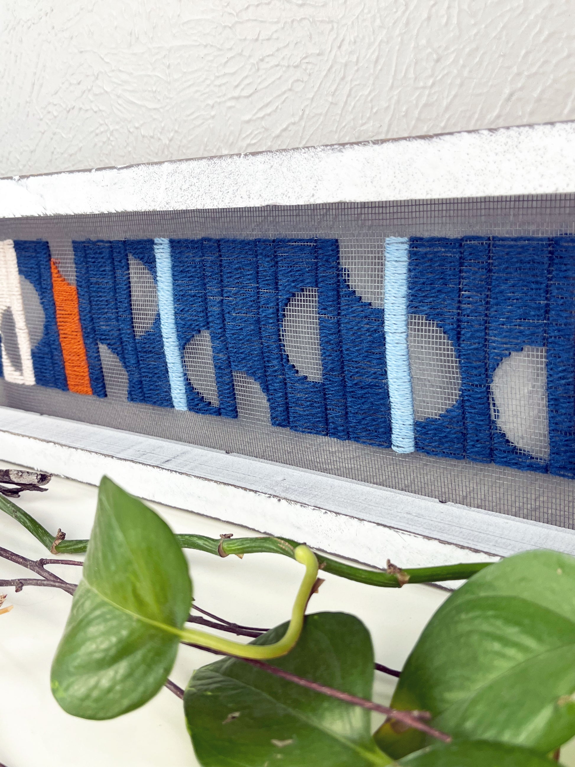 close up view of a window screen framed in a long thick white frame, the screen is hand stitched with columns of midnight blue light blue rust and peach with negative space semi circles, on a white counter with pothos vines and dried sticks around it
