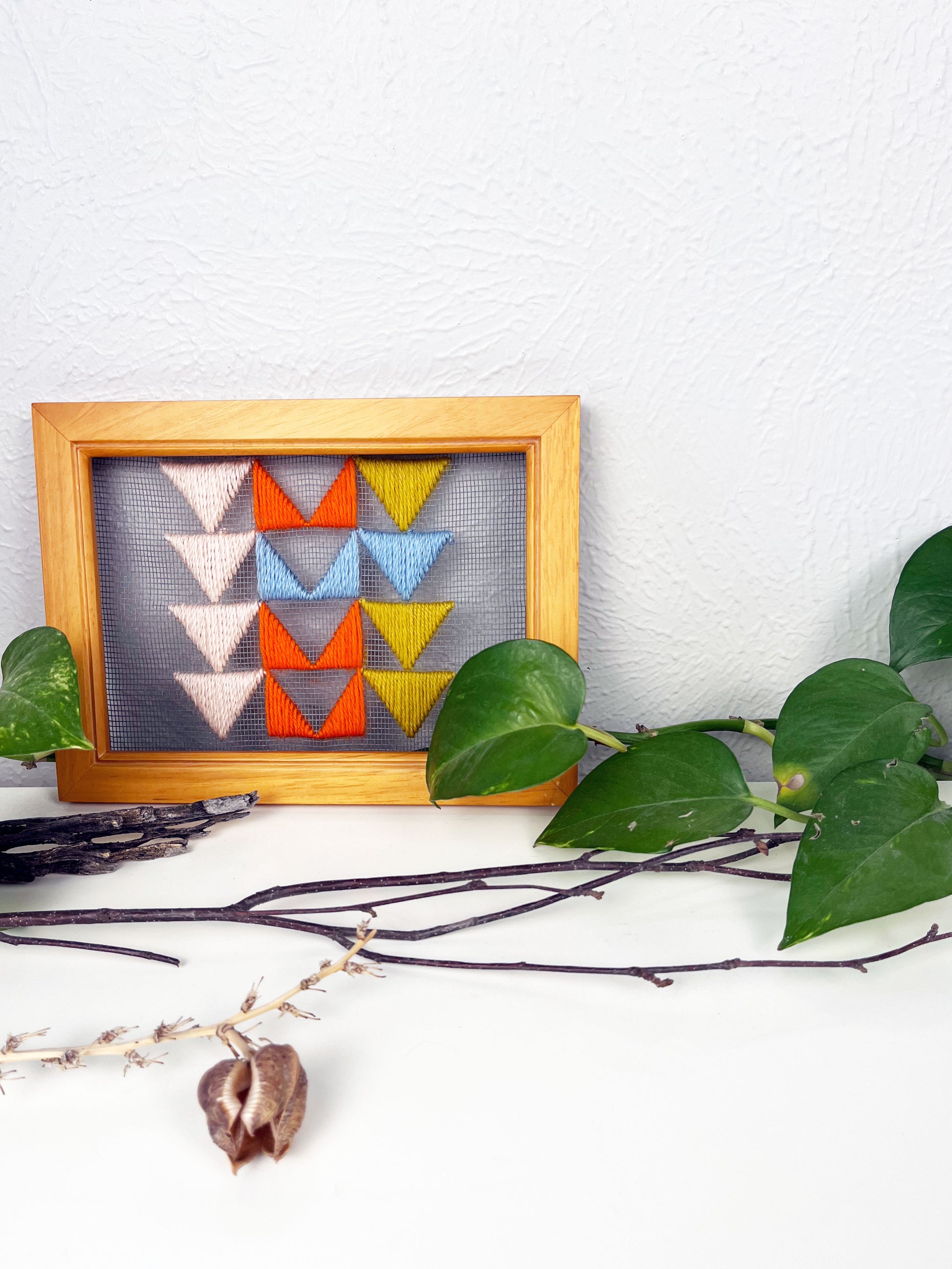 a piece of window screen hand stitched with 3 rows of triangles resembling the flying geese quilt blocks, in neon orange light blue mustard yellow and peach, in a wood frame, on a white counter, surrounded by a pothos vine, dried flowers and sticks