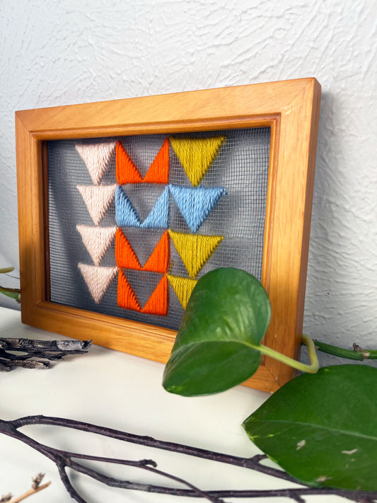 a piece of window screen hand stitched with 3 rows of triangles resembling the flying geese quilt blocks, in neon orange light blue mustard yellow and peach, in a wood frame, on a white counter, surrounded by a pothos vine, dried flowers and sticks
