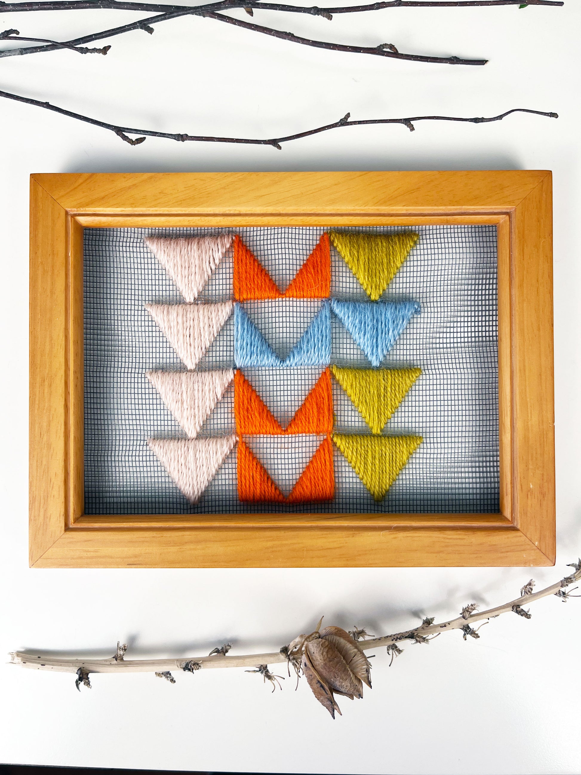 a piece of window screen hand stitched with 3 rows of triangles resembling the flying geese quilt blocks, in neon orange light blue mustard yellow and peach, in a wood frame, on a white counter, surrounded by dried flowers and sticks