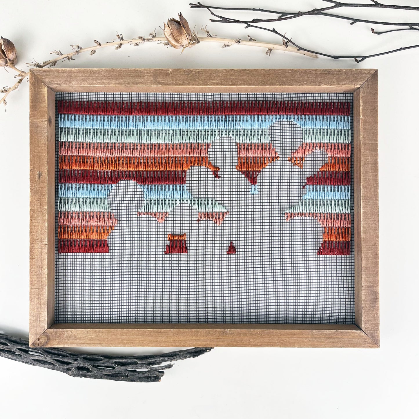 a piece of window screen hand stitched with rows of maroon rusty orange mint green and light blue stitches with an image in negative space of a prickly pear cactus, in a wood frame, on a white counter, with pieces of dried yucca pods and cholla cactus wood around it