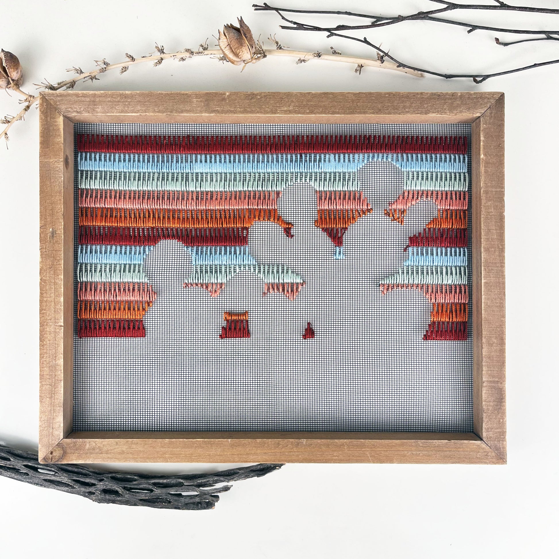 a piece of window screen hand stitched with rows of maroon rusty orange mint green and light blue stitches with an image in negative space of a prickly pear cactus, in a wood frame, on a white counter, with pieces of dried yucca pods and cholla cactus wood around it