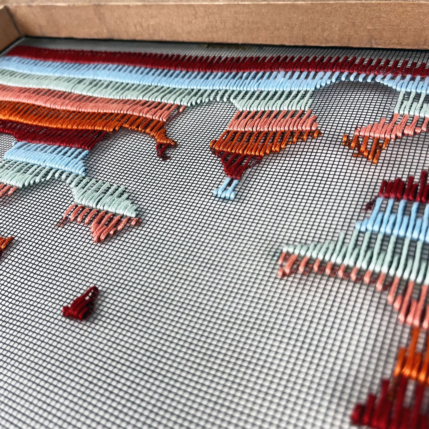 close up angled view of a piece of window screen hand stitched with rows of maroon rusty orange mint green and light blue stitches with an image in negative space of a prickly pear cactus, in a  wood frame, on a white counter