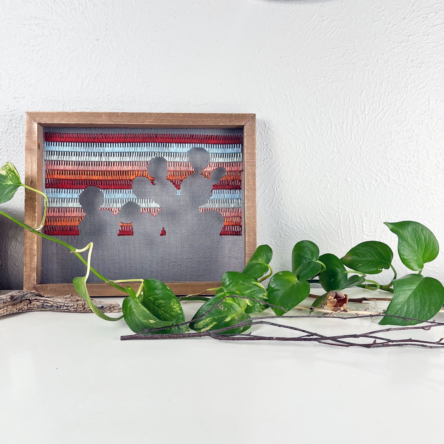 a piece of window screen hand stitched with rows of maroon rusty orange mint green and light blue stitches with an image in negative space of a prickly pear cactus, in a wood frame, on a white counter, with a pothos vine around it