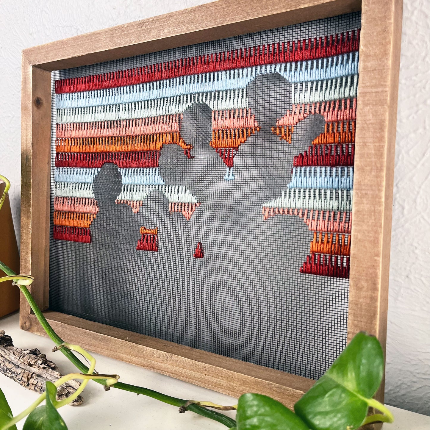 close up angled view of a piece of window screen hand stitched with rows of maroon rusty orange mint green and light blue stitches with an image in negative space of a prickly pear cactus, in a wood frame, on a white counter, with a pothos vine around it