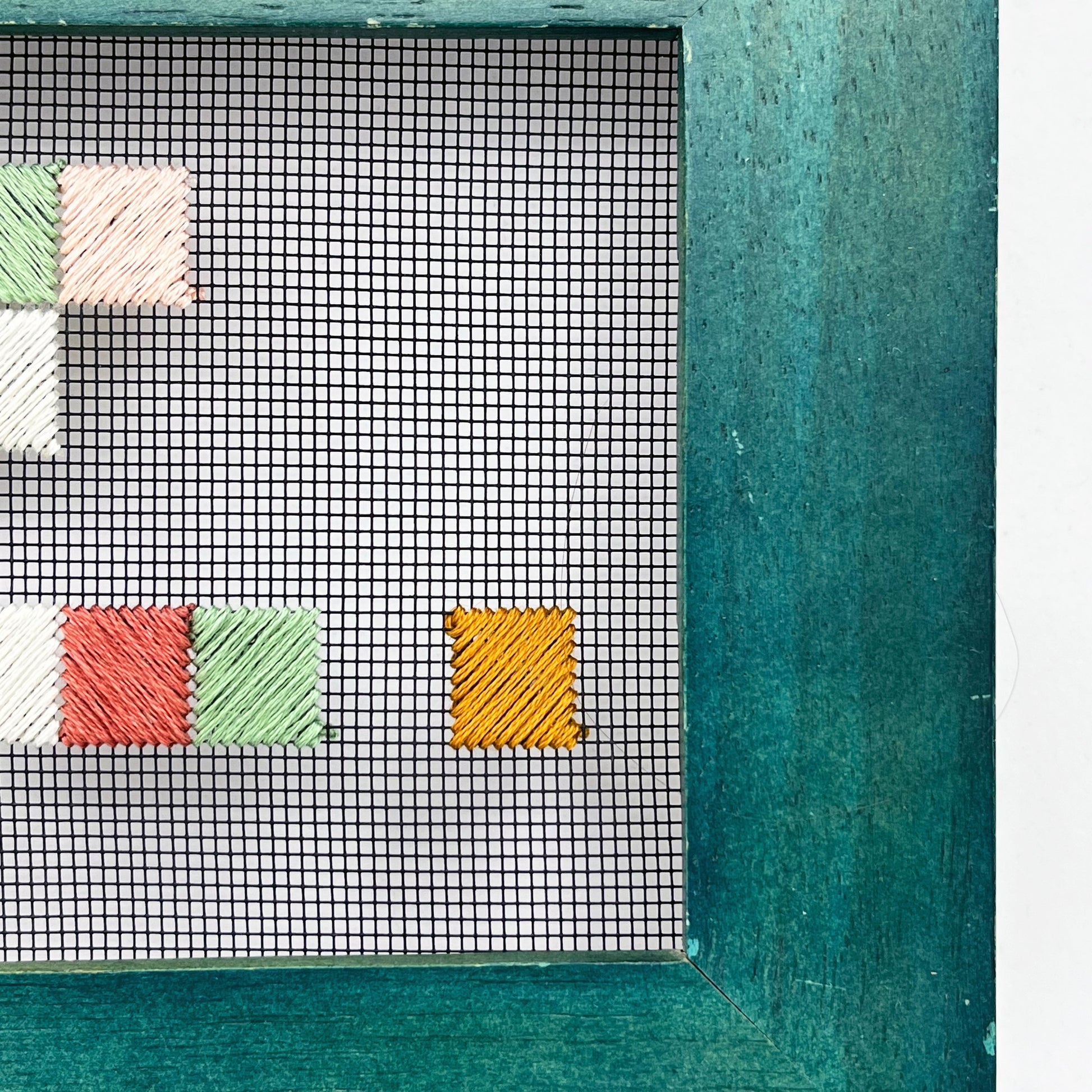 a very close up abstract view of the corner of a piece of window screen hand stitched with an abstract design made of squares with diagonal stitches, in greens peaches and mustard, in a wide green frame