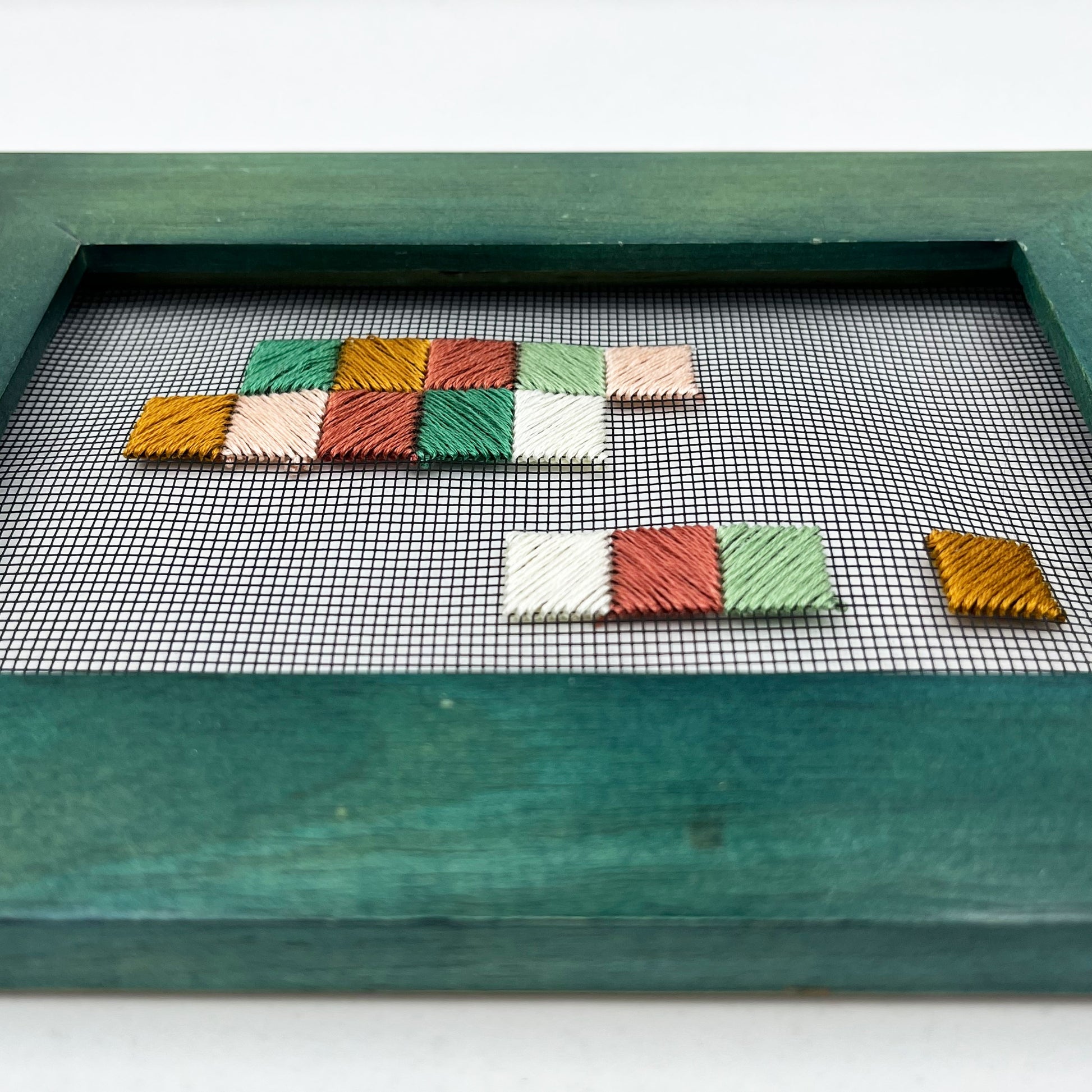 a close up angled view of a piece of window screen hand stitched with an abstract design made of squares with diagonal stitches, in greens peaches and mustard, in a wide green frame