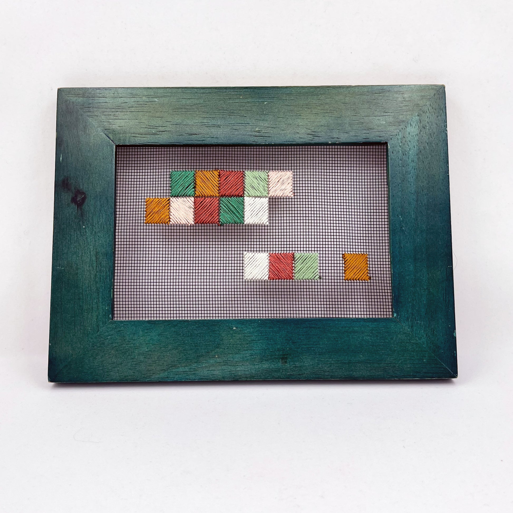 a piece of window screen hand stitched with an abstract design made of squares with diagonal stitches, in greens peaches and mustard, in a wide green frame, on a white background