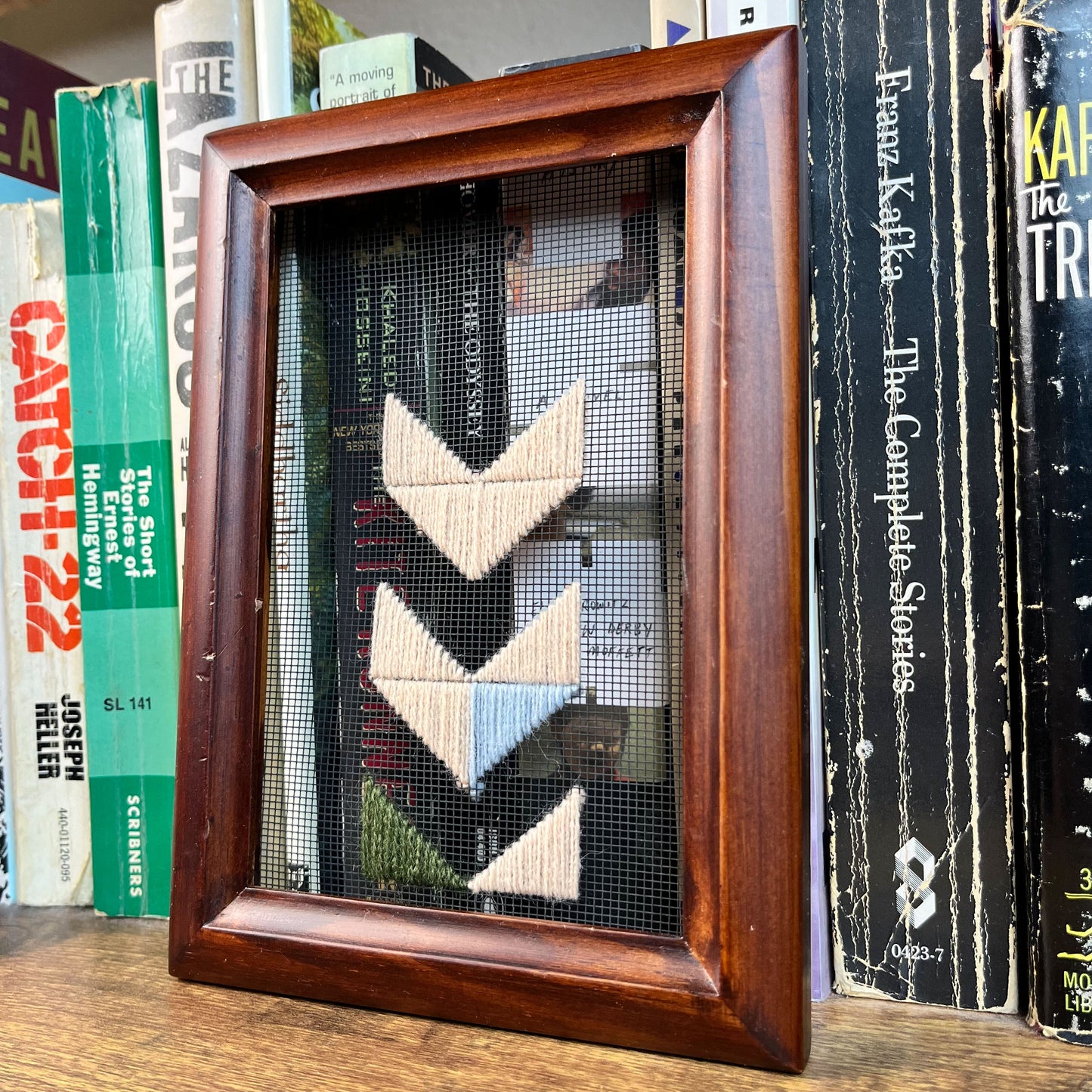 a piece of window screen hand stitched with triangles forming large chevrons centered on the screen, running off the bottom, mostly in peach with some light blue and olive green, in a wood frame, in front of a row of books
