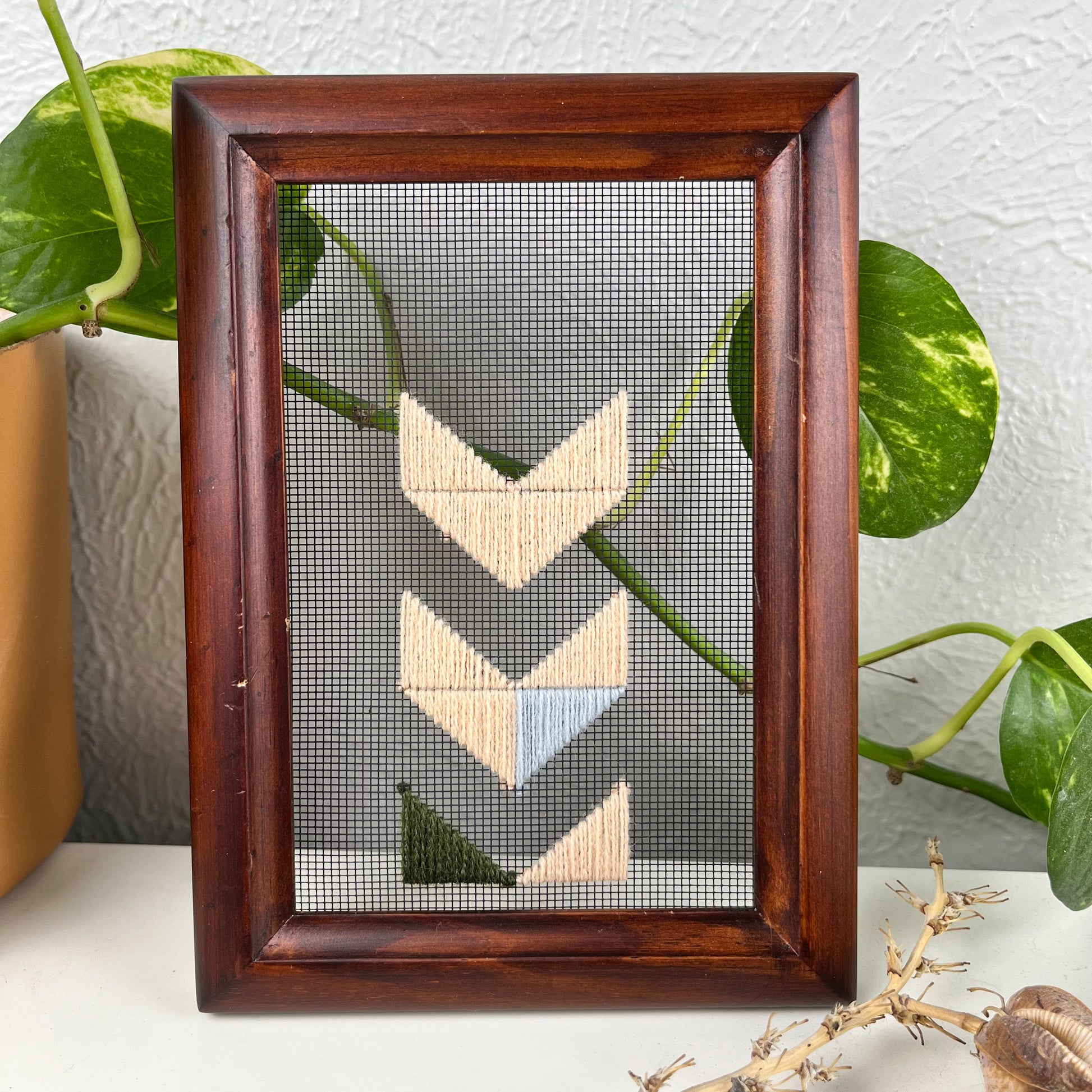 a piece of window screen hand stitched with triangles forming large chevrons centered on the screen, running off the bottom, mostly in peach with some light blue and olive green, in a wood frame, on a white counter, with a pothos behind it