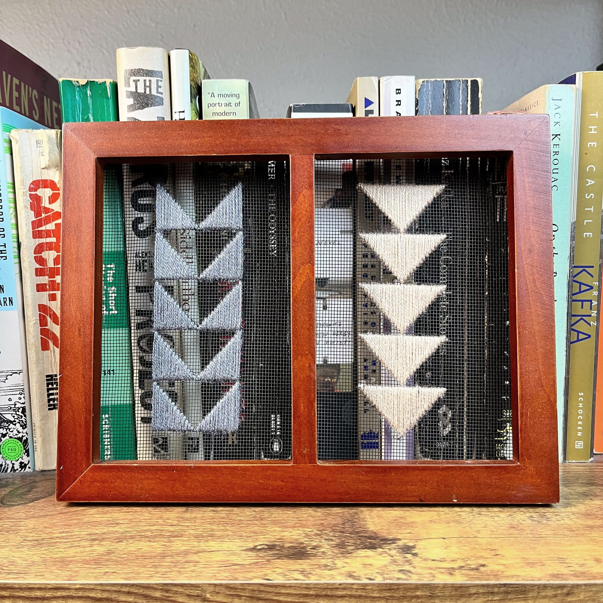 a piece of window screen hand stitched with two rows of triangles resembling the flying geese quilt blocks, in grey and peach, in a double wood frame, in front of a row of books