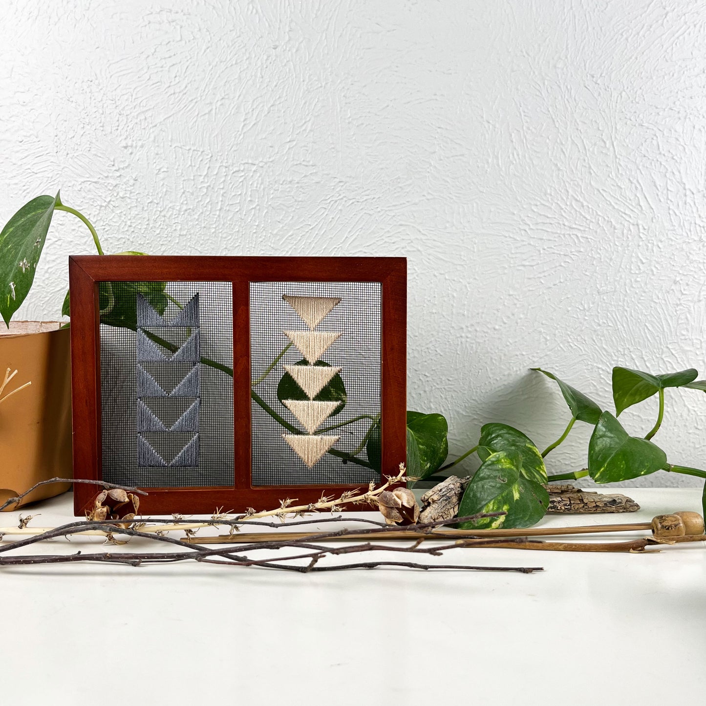 a piece of window screen hand stitched with two rows of triangles resembling the flying geese quilt blocks, in grey and peach, in a double wood frame, on a white counter, surrounded by a pothos vine, dried flowers and sticks