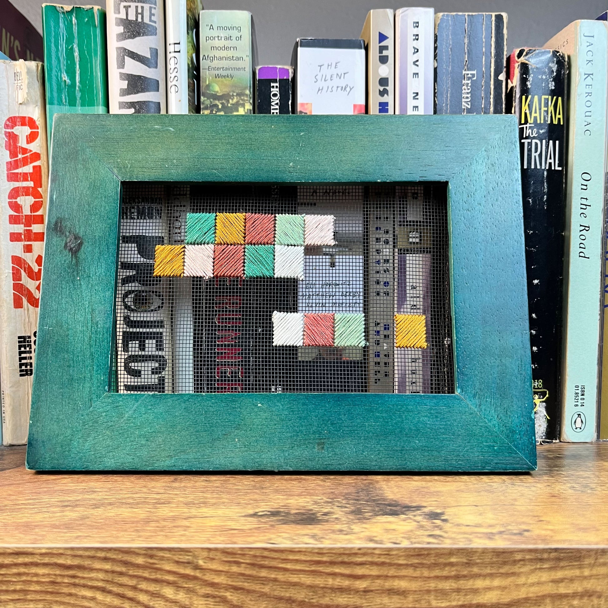 a piece of window screen hand stitched with an abstract design made of squares with diagonal stitches, in greens peaches and mustard, in a wide green frame, resting on a bookshelf in front of a row of books