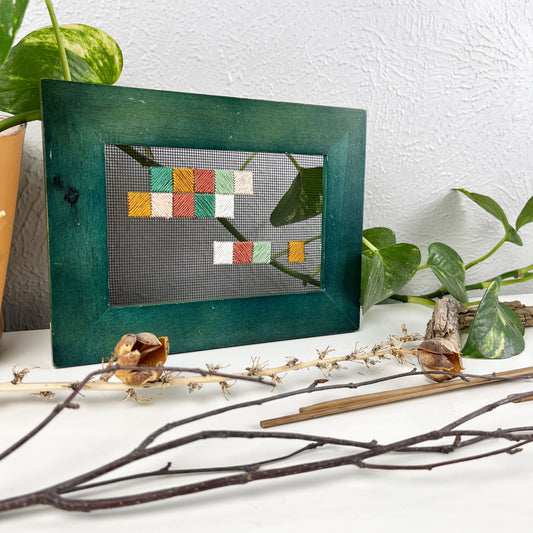 a piece of window screen hand stitched with an abstract design made of squares with diagonal stitches, in greens peaches and mustard, in a wide green frame, on a white counter, surrounded by a pothos vine, dried flowers and sticks
