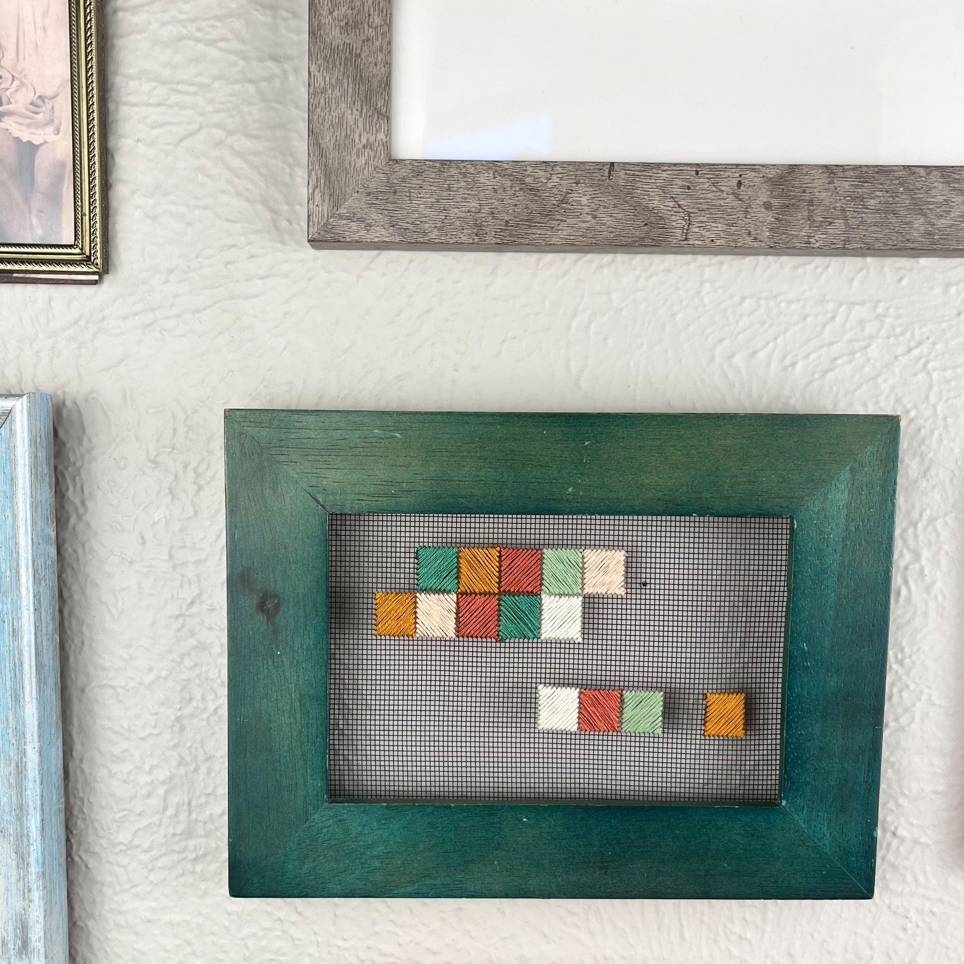 a piece of window screen hand stitched with an abstract design made of squares with diagonal stitches, in greens peaches and mustard, in a wide green frame, on a wall with hints of other frames around it