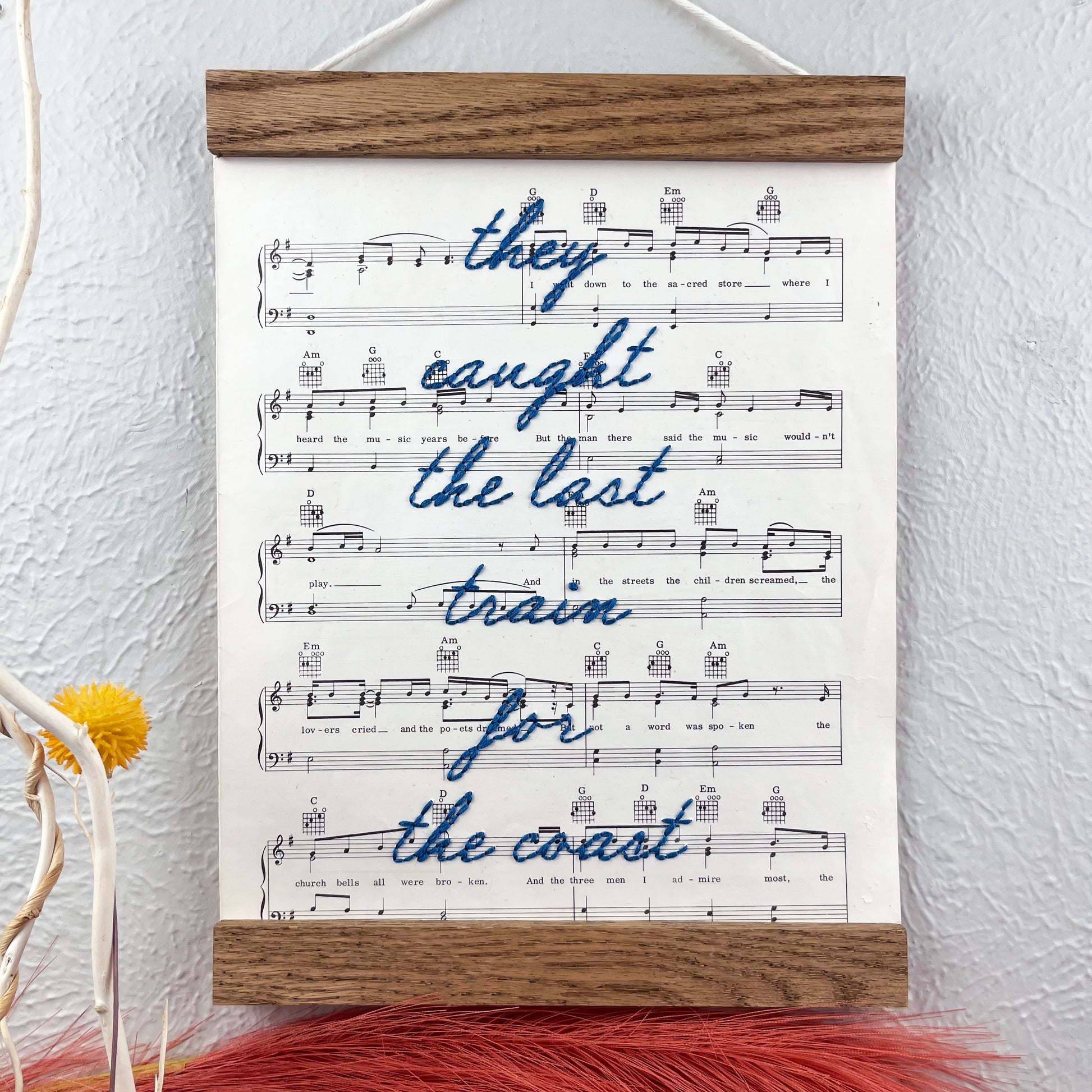 sheet music for the song American Pie by Don McLean hanging in a magnetic wood frame with the words they caught the last train for the coast hand embroidered over it in blue colored thread in cursive hanging on a wall near other framed pictures art and decorative branches